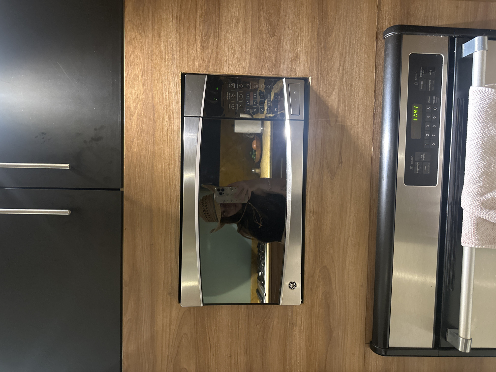 The media file captures a close-up view of a modern kitchen interior, primarily focusing on two built-in appliances and surrounding cabinetry. On the left are dark, possibly matte black, kitchen cabinets with silver horizontal bar handles. To the right, a warm-toned wooden veneer wall separates these cabinets from a built-in microwave and an adjacent stainless steel appliance, which appears to be a refrigerator or oven.
The central subject is a GE brand microwave, integrated into the wooden cabinetry. Its dark, reflective door and control panel feature silver trim. A significant detail is the clear reflection in the microwave's door, showing a person wearing a straw hat and holding a smartphone, actively taking the photograph. This suggests the activity taking place is photography within a kitchen setting. The microwave's control panel displays numbers 1 through 9 and 0, along with various function icons and some text.
To the right of the microwave, another large stainless steel appliance is visible, possibly an oven or refrigerator, with a digital display showing "11:41," indicating the time of day. A white, textured towel hangs from its long, cylindrical handle. The control panel of this appliance features Spanish text, including "TIEMPO" (time), "hornear" (bake), "programar" (program), "cronómetro" (stopwatch), "iniciar" (start), "apagar" (turn off), "luz horno" (oven light), "asar" (roast), "temperatura" (temperature), and "HORNO" (oven).
The overall setting is a well-lit kitchen, with a clean and contemporary design, as suggested by the sleek appliances and combination of dark and wooden finishes. The location context indicates this scene is in Mexico City, Mexico.