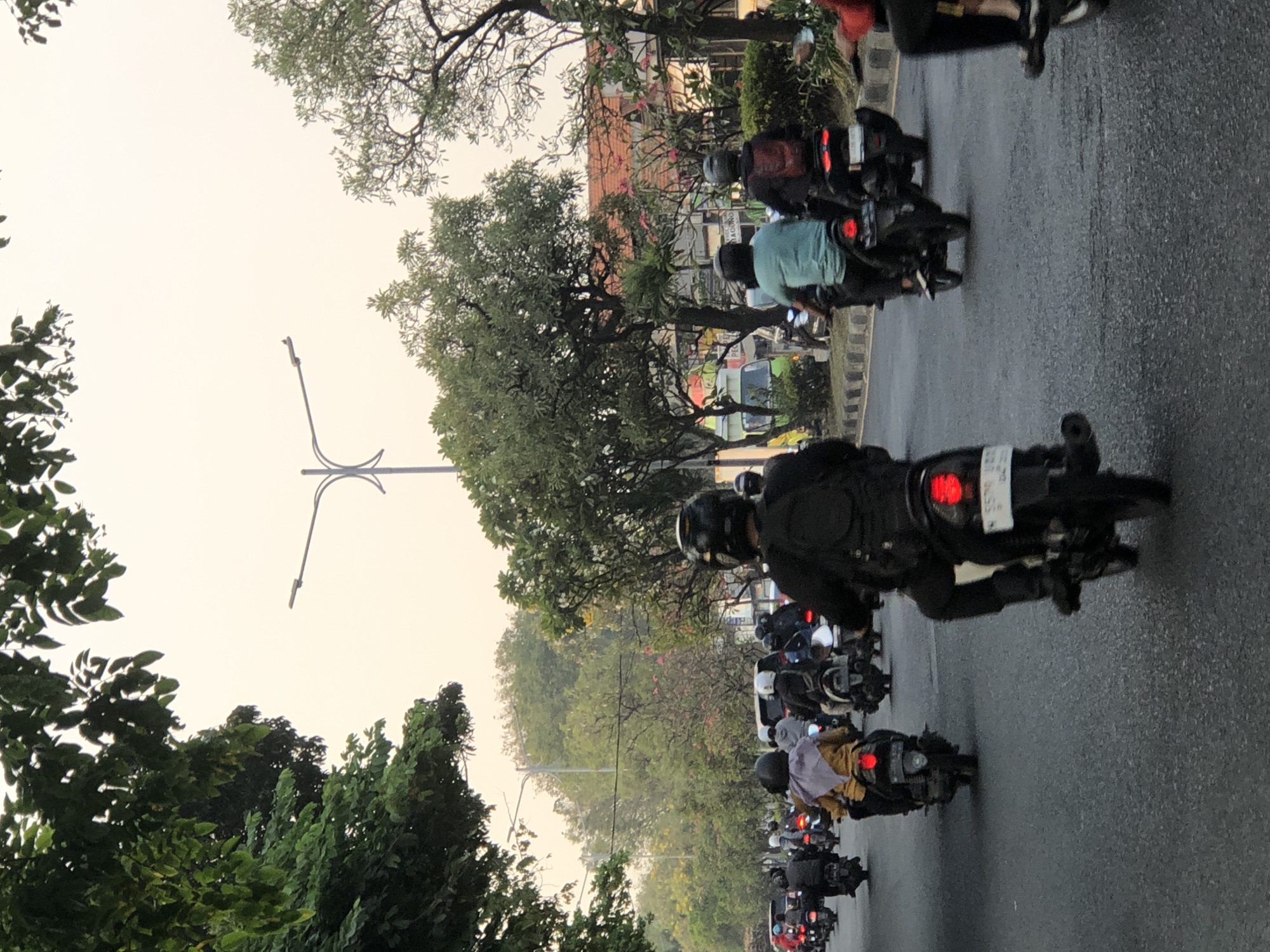 The image depicts a street scene in Sumorame, Indonesia, with a procession of motorcycles traveling along a road. The perspective is from behind the riders, looking forward. Several individuals are visible on the motorcycles, most wearing helmets. The environment includes trees lining the street, a lamppost, and buildings in the background. The sky is overcast and light. The road surface is asphalt. The motorcycles are diverse in model and color. Some riders are wearing backpacks and casual clothing. The overall atmosphere suggests traffic flow in a moderately populated urban or semi-urban area.
