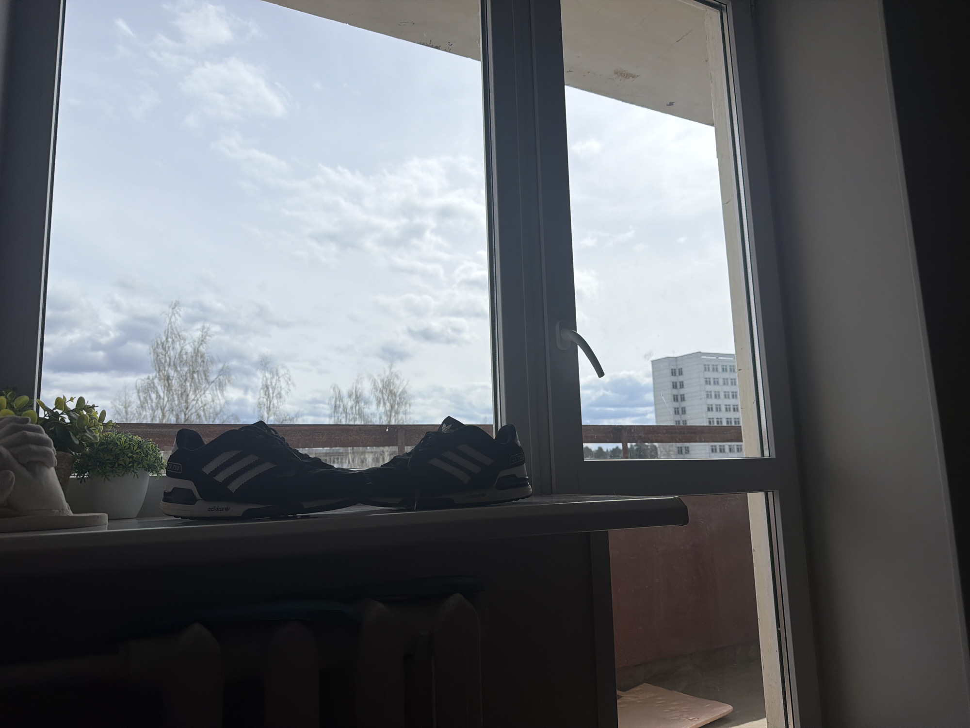 The image captures an indoor perspective looking out through a large window, framing a view of a cloudy sky and distant landscape. In the foreground, on a wide windowsill, two dark sneakers are positioned, with one clearly displaying the "adidas" brand name on its sole and "ZX 750" partially visible on its side. To the left of the sneakers, a small potted plant with green foliage and a white decorative object, possibly a small statuette fragment, are also on the sill. Below the windowsill, a dark radiator is partially visible, indicating an indoor heating system.
The window frames, dark and modern, divide the external view. Beyond the window, a bright, overcast sky with soft clouds occupies the upper part of the scene. Below the horizon, a line of bare trees, suggesting late winter or early spring, stretches across the middle ground. To the far right, a tall, multi-story white building with many windows stands prominently, a common sight in urban landscapes like Chaikovsky, Russia. Below the trees and distant building, an outdoor balcony area with a reddish-brown wall and a wooden railing is discernible. The indoor area is in shadow, creating a contrast with the brighter outdoor light, indicating it is daytime. The scene is static, capturing a quiet moment looking out onto a typical urban-natural environment.