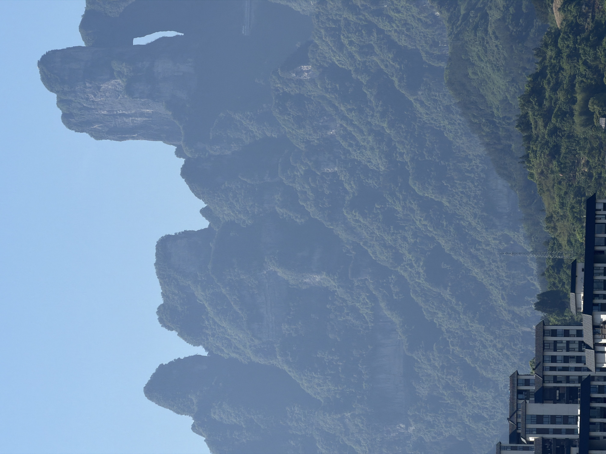 A daytime shot depicts the Tianmen Mountain in Zhangjiajie, China. The mountain, partially obscured by mist, dominates the frame with its steep, vegetation-covered slopes and unique natural archway near the summit, known as Tianmen Cave. Modern buildings are present in the foreground, providing a contrasting element to the natural landscape. An electrical transmission tower is visible mid-frame.