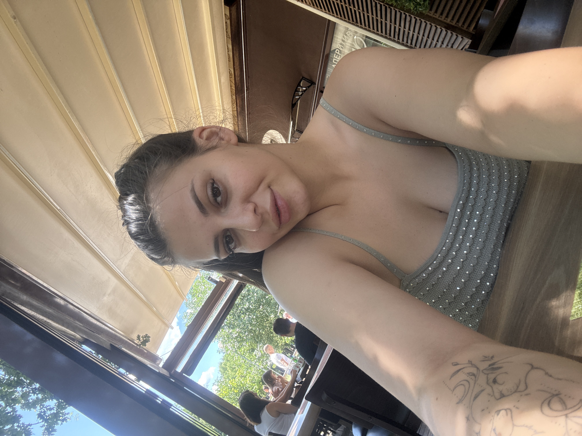 A young woman with dark hair pulled back in a ponytail is seated at an outdoor table, posing for a selfie. She is wearing a grey, sleeveless top adorned with small, reflective embellishments. Her left arm is visible, featuring a tattoo of a cat and other illustrative elements. In the background, through an open-air structure with a retractable awning, trees and other patrons can be seen. The overall setting appears to be a cafe or restaurant with an outdoor seating area. The lighting suggests it is daytime, likely sunny. The architectural style of the awning and the presence of greenery suggest a European location, potentially Plovdiv, Bulgaria, given the hint provided. The woman's gaze is directed towards the camera, and she has a slight smile. The table she is sitting at appears to be made of wood.