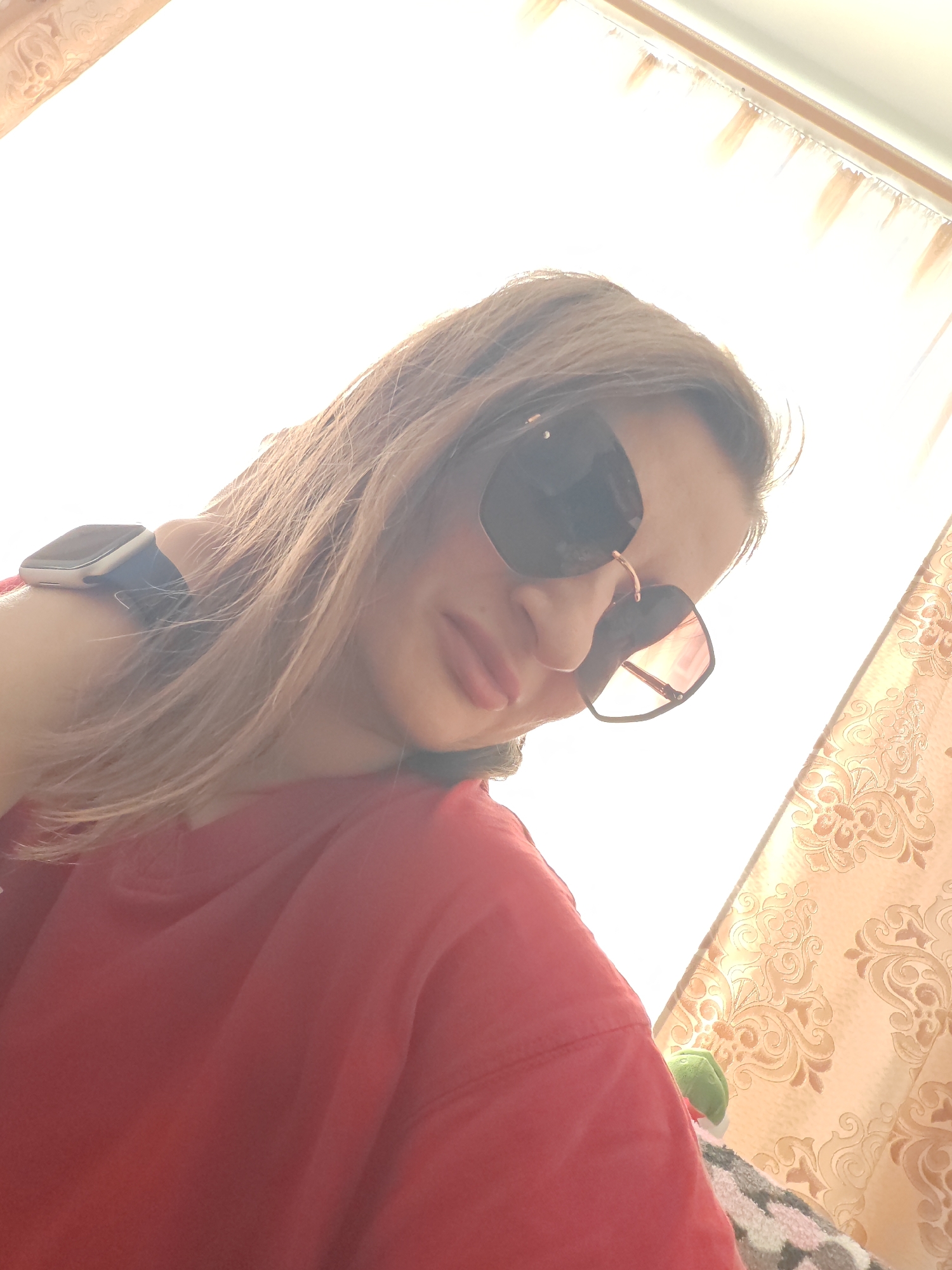A woman with blonde hair and sunglasses is seen from a low angle, looking down. She is wearing a red shirt and a watch on her left wrist. She is positioned in front of a window with curtains, and a stuffed toy is visible on her right. The scene is illuminated by bright light from above. The background shows a patterned curtain and a window with fringe.