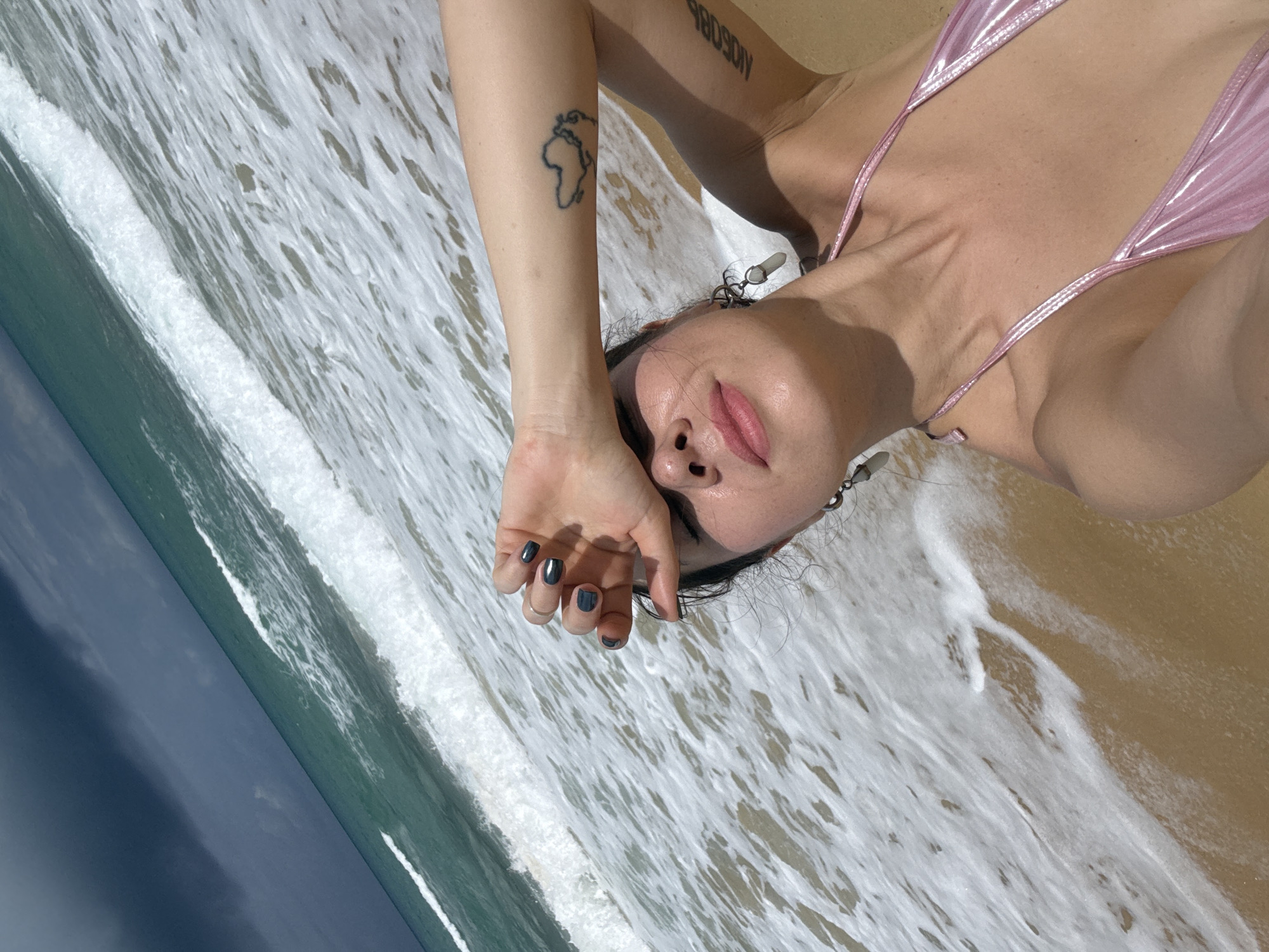 A woman is taking a selfie on a beach. She is wearing a pink bikini and has her hand over her eyes, shielding them from the sun. The ocean waves are breaking on the shore behind her, and the sky is dark with storm clouds. The location is the city of Karon, Thailand.