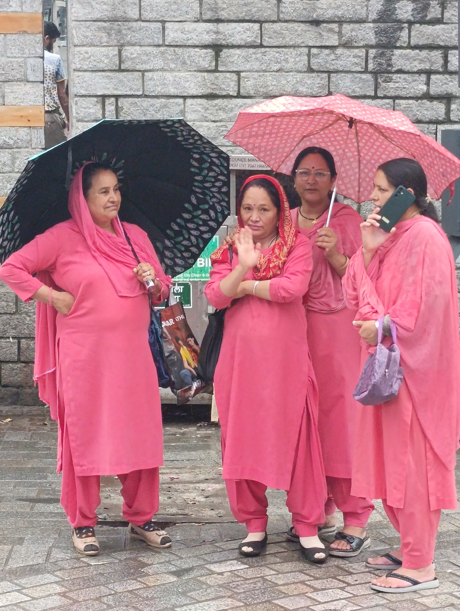 Four women, dressed in matching bright pink salwar kameez outfits, stand together outdoors on a paved walkway in Manali, India. Two women are holding open umbrellas, one black with a white feather-like pattern and the other pink with a repeating circular design. The woman on the far left holds the black umbrella and has her left hand on her hip. The woman next to her, in the center of the frame, waves with her right hand and wears a red and gold patterned scarf around her head. The third woman from the left, holding the pink umbrella, has her arms crossed. The woman on the far right is talking on a dark green smartphone and carries a small, textured purple handbag in her left hand. Behind the women, a brick wall forms the background, with a glimpse of a man in the background to the left, partially obscured by a doorway. The ground appears wet, suggesting it may have recently rained.