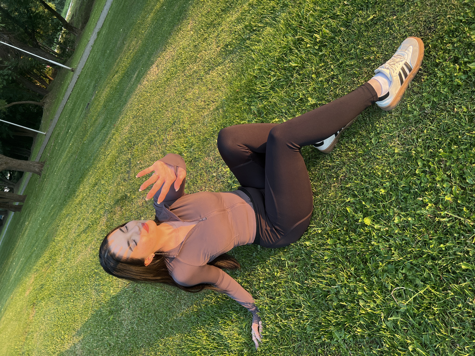 A young woman with long dark hair is sitting on a green grassy lawn, with the sun shining and casting shadows. She is wearing a brown athletic top with a zipper and black leggings. Her eyes are closed and she is holding up her hand to block the sun. She is wearing white sneakers with black stripes. The background shows a park with trees and a pathway. The location is the city of Marushkino, Russia.
