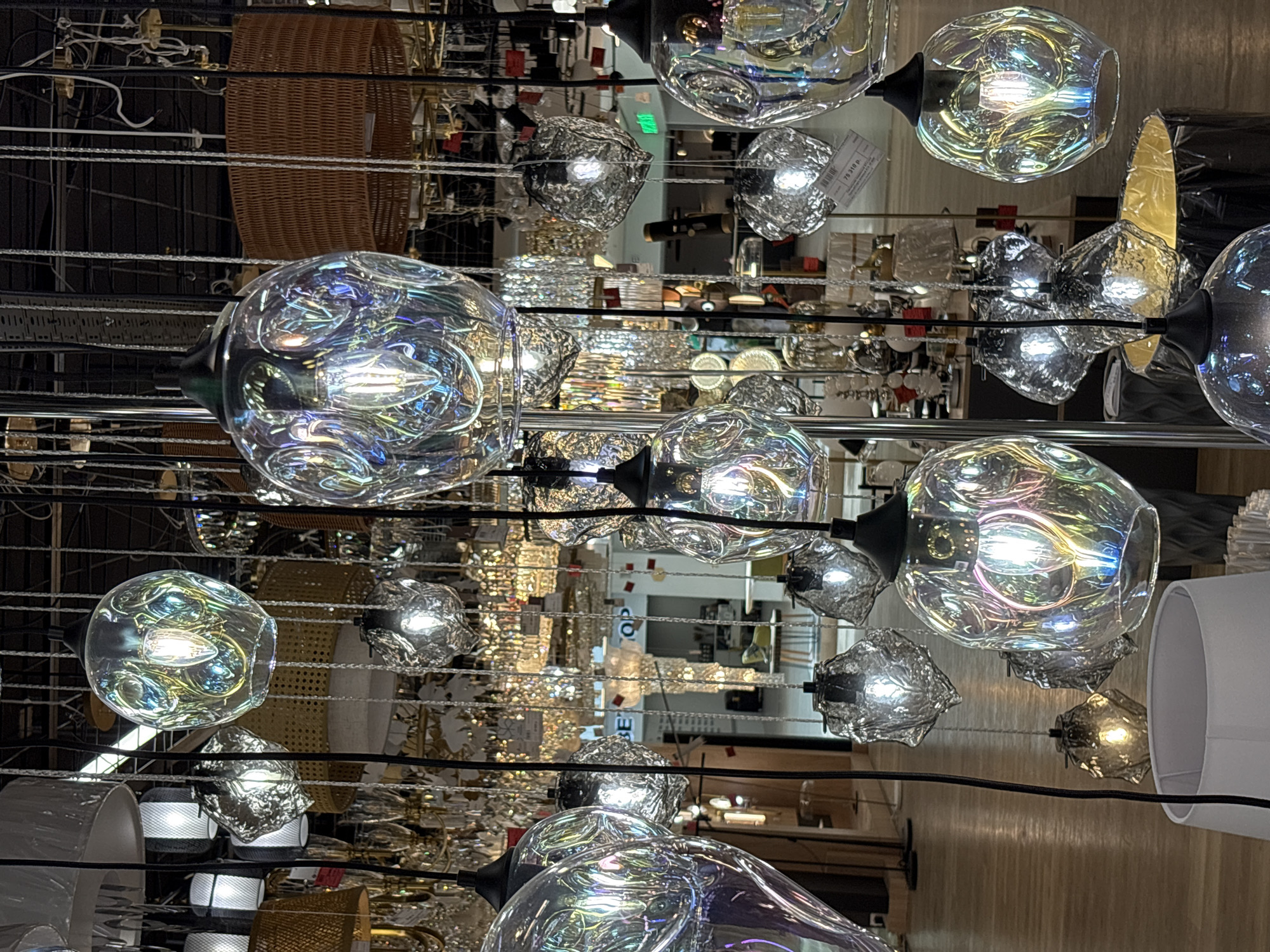 The image depicts an indoor display of various decorative light fixtures, predominantly pendant lamps, in a retail setting. The lighting fixtures feature an assortment of glass shades, some with iridescent or textured finishes, and others with a more hammered or crumpled appearance. These are suspended by wires or cords against a dark background, likely a ceiling. Behind the primary display, numerous other light fixtures are visible, creating a layered and somewhat cluttered effect. These include chandeliers, lampshades, and what appear to be woven basket-like elements. The floor below is made of polished wood, reflecting some of the light. In the background, glimpses of store aisles, display shelves, and possibly other retail elements are discernible, suggesting a lighting showroom or department store. The overall impression is one of a well-lit and varied merchandise display. No people are present in the frame. This scene is consistent with a retail environment in Saint Petersburg, Russia, specifically a lighting store within a commercial complex or a dedicated showroom.