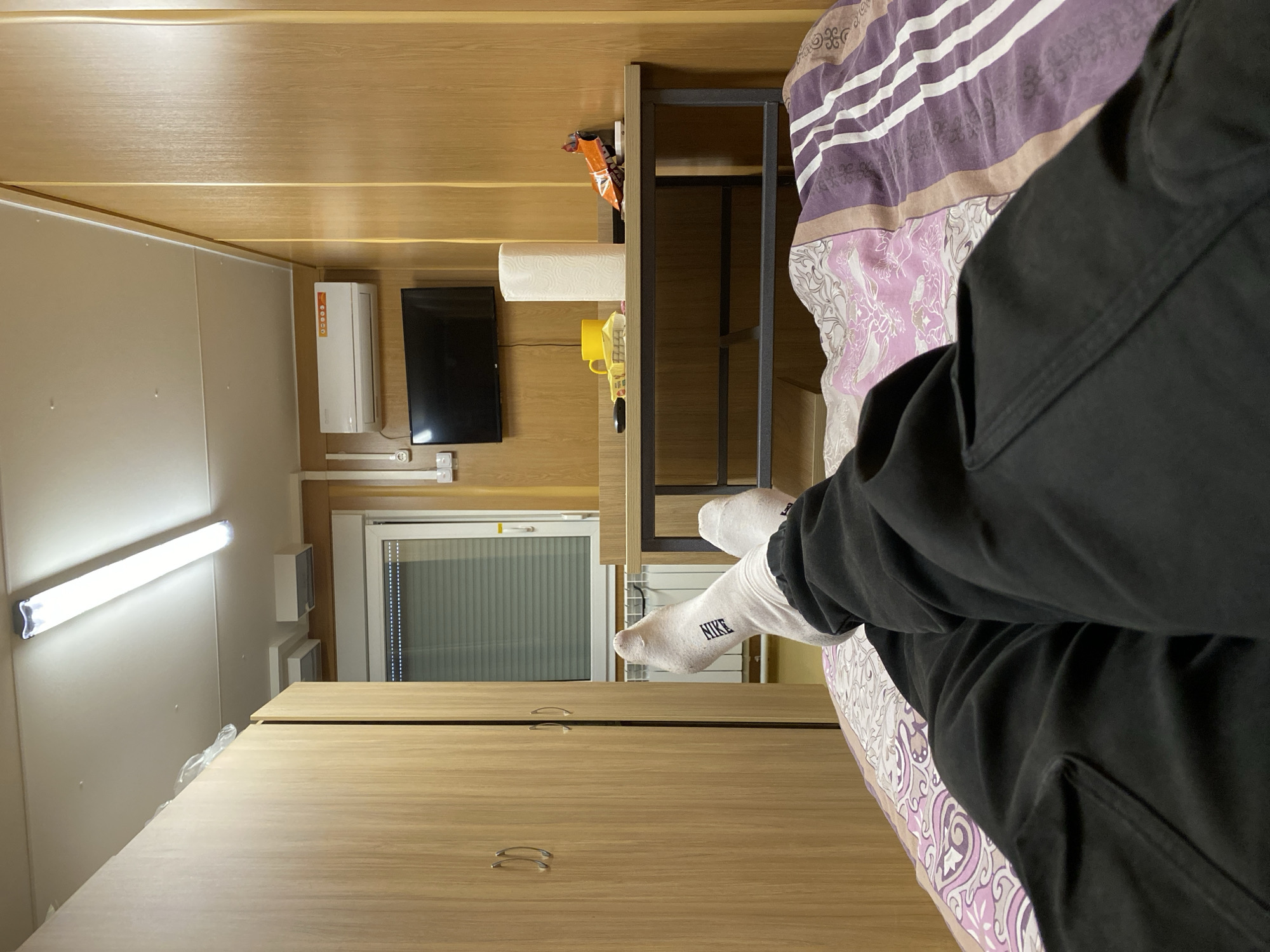 The image captures an indoor scene from a first-person perspective, looking from a bed towards the opposite wall of a room. The viewer's lower legs and feet are visible in the foreground on the right side of the frame, clad in black pants or sweatpants and white socks. The left sock clearly displays the brand "NIKE". The bed is partially visible, covered with a duvet featuring a purple, white, and brown/beige patterned design.
The room appears to be compact, possibly a dormitory or small apartment, with light-colored wood paneling covering the walls and ceiling. Directly ahead, at the foot of the bed, there is a wooden cabinet or desk with two silver-colored handles. Above this furniture, a window with interior blinds or a screen is visible, suggesting a view to the outside.
Mounted on the wall above the window are a white air conditioning unit and a black flat-screen television. Below the television, a white power strip or set of outlets is installed. To the left of the window, a long, bright white fluorescent light fixture provides illumination. To the right of the TV, a dark metal shelving unit is present. On a shelf above the television, items such as a roll of paper towels, a yellow mug, and several snack packages (one with an orange wrapper) are placed. Electrical wires are visible connecting to the television and power sources. The scene depicts a moment of relaxation in a functional, well-lit room.