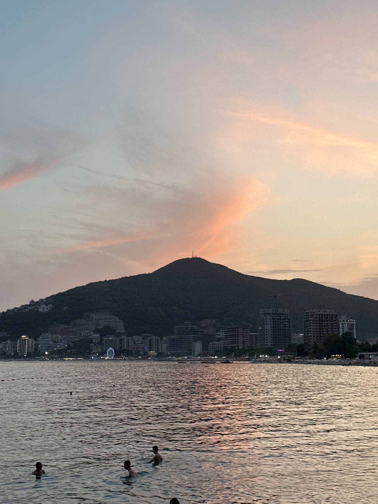 This image captures a serene sunset scene over the city of Budva, Montenegro, likely from its coastline. The sky is painted with soft hues of pink and orange from the setting sun, with wispy clouds streaking across. Dominating the background is a dark, tree-covered mountain, its silhouette sharp against the colorful sky. Atop the mountain, a small antenna or communication tower is visible.
Nestled on the slopes of the mountain and along the coast is the cityscape of Budva. Numerous buildings, ranging from older, smaller structures to modern, multi-story hotels and apartment complexes, are visible. Some buildings appear to be under construction, with cranes present. A Ferris wheel is discernible in the distance, indicating a recreational area or amusement park within the city.
In the foreground, the Adriatic Sea stretches out, its surface rippling gently. The water reflects the soft colors of the sky, creating a shimmering effect. Several individuals are present in the water, with some appearing to be swimming or wading. Their bodies are partially submerged, and their presence suggests a beach or swimming area. Beach umbrellas and seating are also visible along the shoreline in the midground, implying a populated tourist area. The overall atmosphere is tranquil and picturesque, typical of a Mediterranean coastal town at dusk.