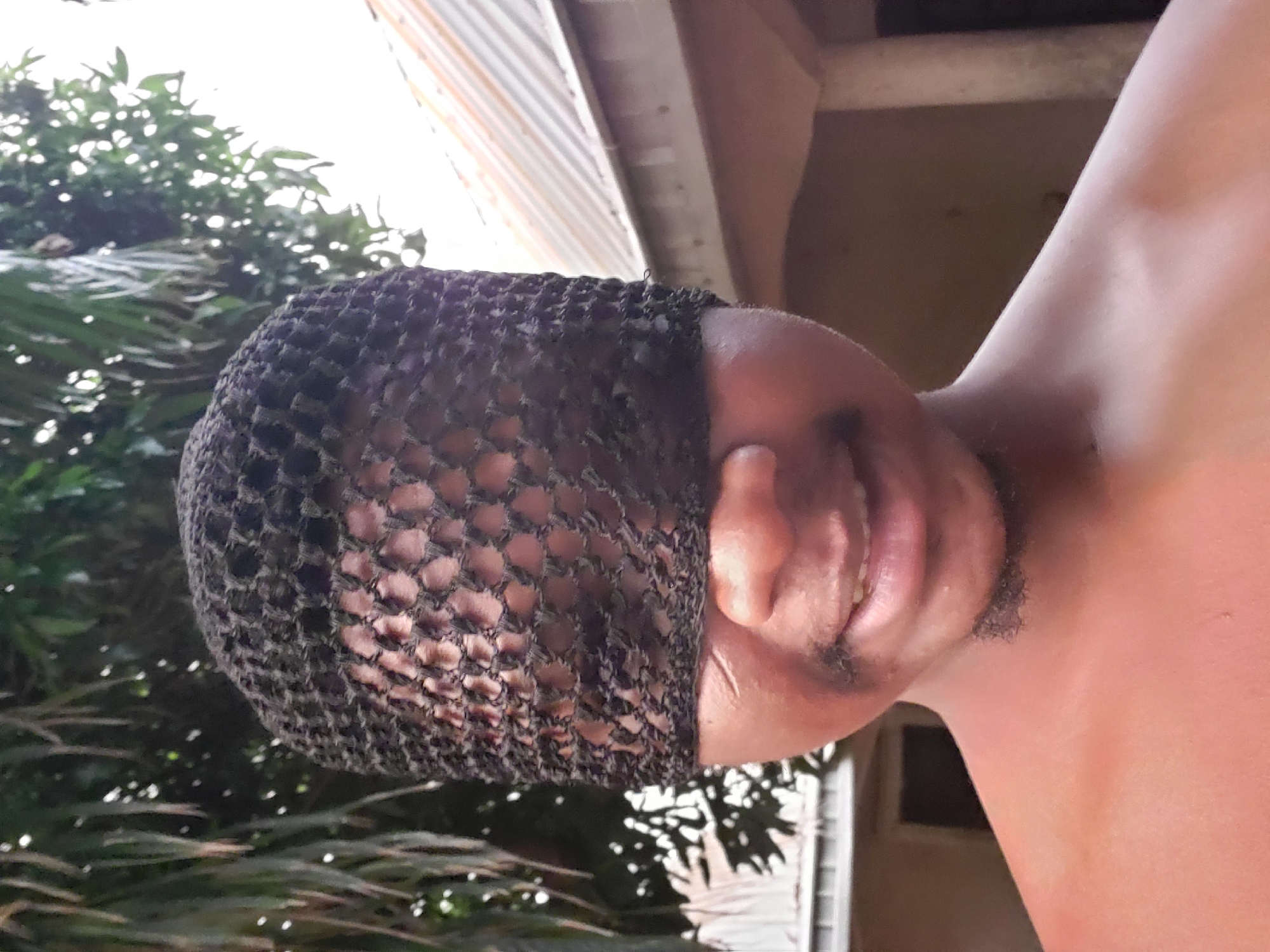 An African male is wearing a black hair net. The image may have been taken in Mararaba, Nigeria, given the user's request. Vegetation is visible. The male's left side of the face and neck are showing. The location appears to be outdoors.