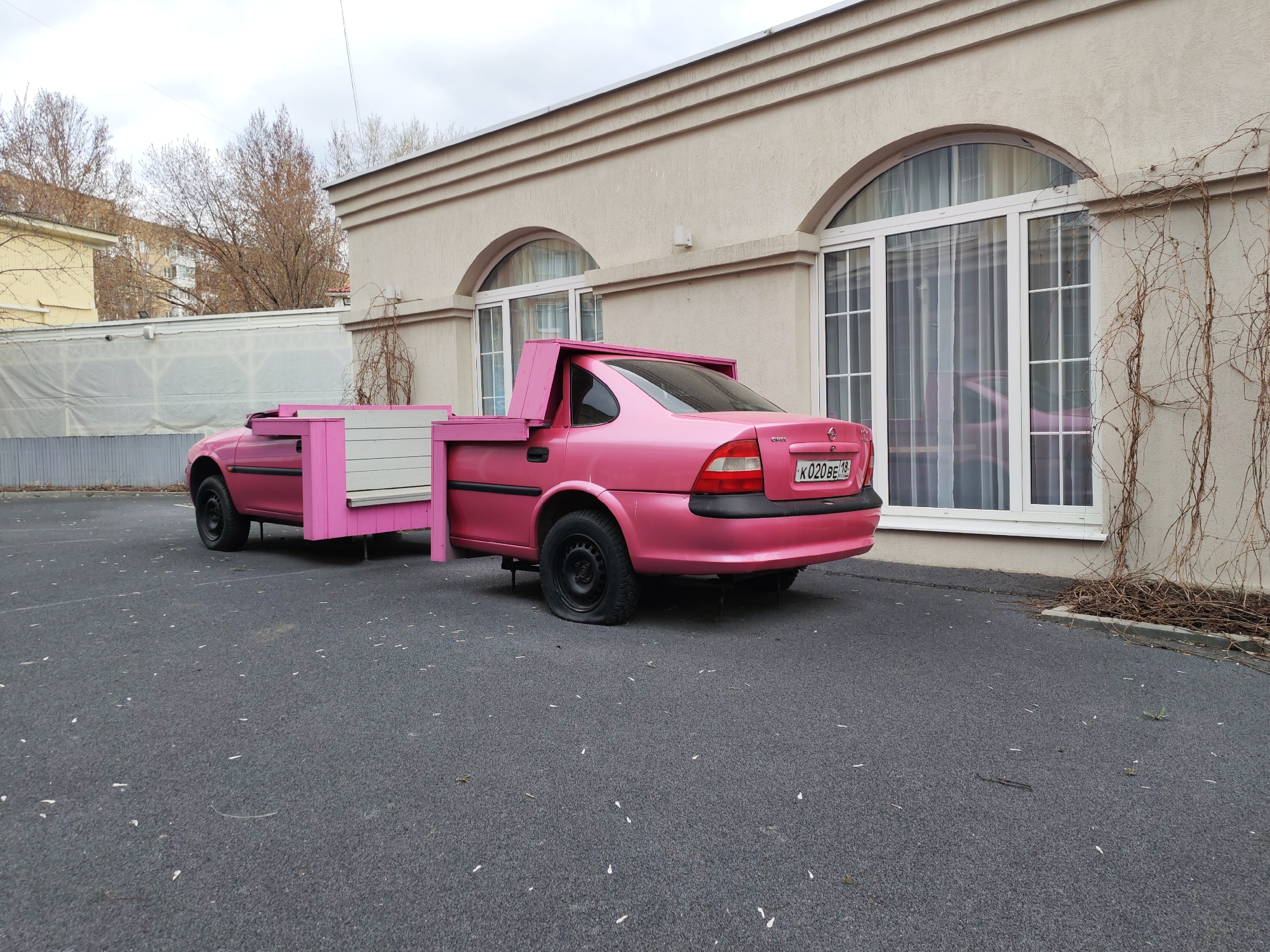 The image shows a pink Opel Vectra car that has been modified into a planter. The rear of the car has been removed and replaced with a wooden box, also painted pink, for holding plants. The car has a Russian license plate, "K 020 BE 18." The rear passenger-side tire is flat. The car is parked on a dark grey asphalt surface in front of a light beige building with arched windows. There are trees and another building visible in the background. The location is likely Izhevsk, Russia, based on the license plate region code (18).