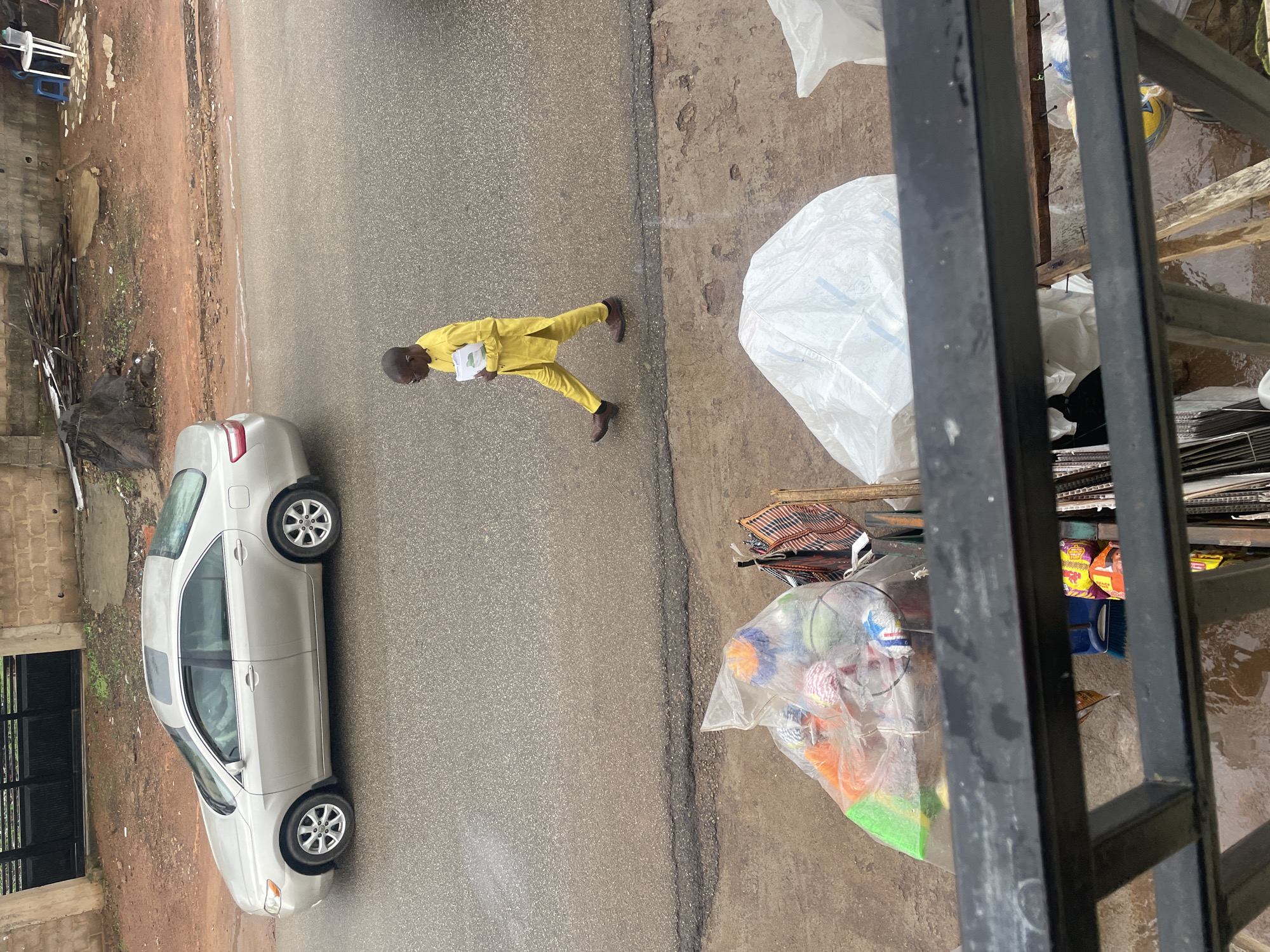 A man in a yellow traditional outfit walks across a wet asphalt road in Gura Top, Nigeria. He is carrying a stack of papers. To his left, a silver sedan is parked on the road. In the foreground, a black metal railing obscures a view of the street, where various items are displayed, including a clear plastic bag filled with colorful balls or toys, and other assorted goods wrapped in plastic. The background features a brick wall and a fence. The overall scene suggests an outdoor market or roadside setting, likely during or after rainfall.