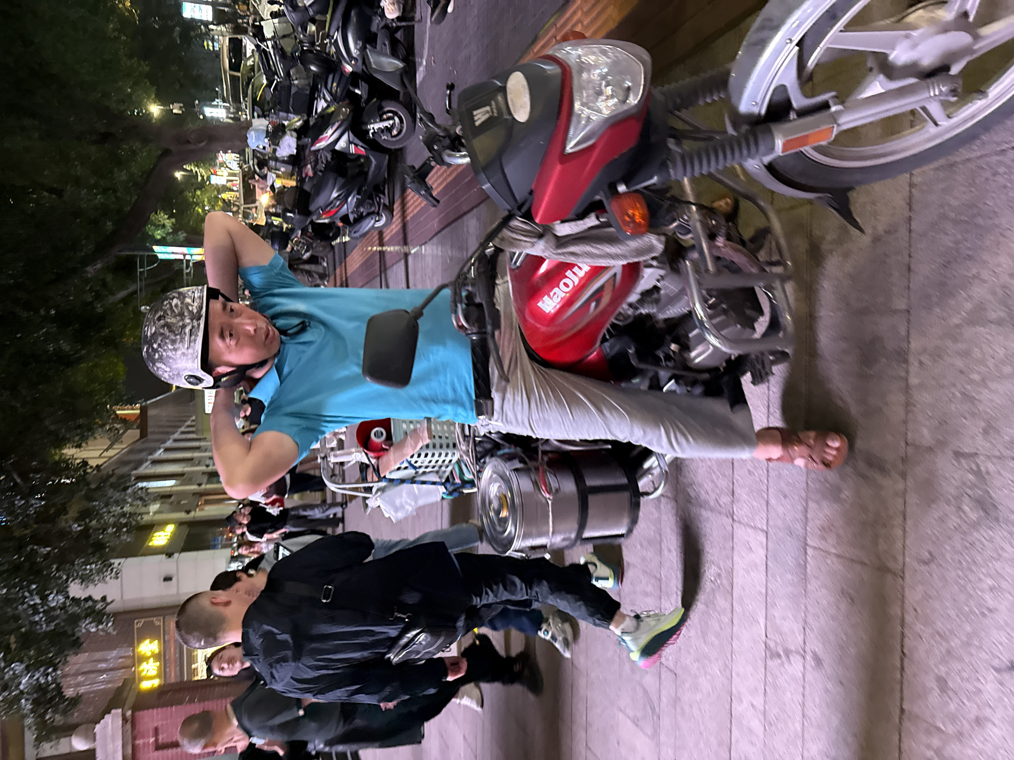 The photograph captures a night scene in an urban environment, likely a street or busy pedestrian area in Yuzhong District, China. The primary subject is a man, appearing to be in his 30s or 40s, wearing a light blue short-sleeved shirt, light-colored pants, and sandals. He sits astride a red motorcycle or scooter, with his hands clasped behind his head, looking towards the camera with a neutral expression. He wears a grey and black camouflage-patterned helmet.
The motorcycle itself is red with silver accents, prominently displaying the brand name "Haojue" (豪爵) on its fuel tank. A black rearview mirror is visible on the left side, and a large silver, cylindrical container, possibly a delivery box, is attached to the rear of the vehicle. The front wheel of the scooter is silver.
In the background, numerous other motorcycles and scooters are densely parked, indicating a common mode of transport or a designated parking zone. Several other people are visible, some walking or standing, including a person in the foreground wearing a black jacket and colorful sneakers. Trees and buildings line the street, illuminated by artificial lighting, suggesting it is nighttime or late evening. On a building in the background, a sign with Chinese characters is visible, including "新街" (Xinjie). The ground is paved with what appears to be concrete or large tiles. The overall setting is bustling, typical of a city street at night.