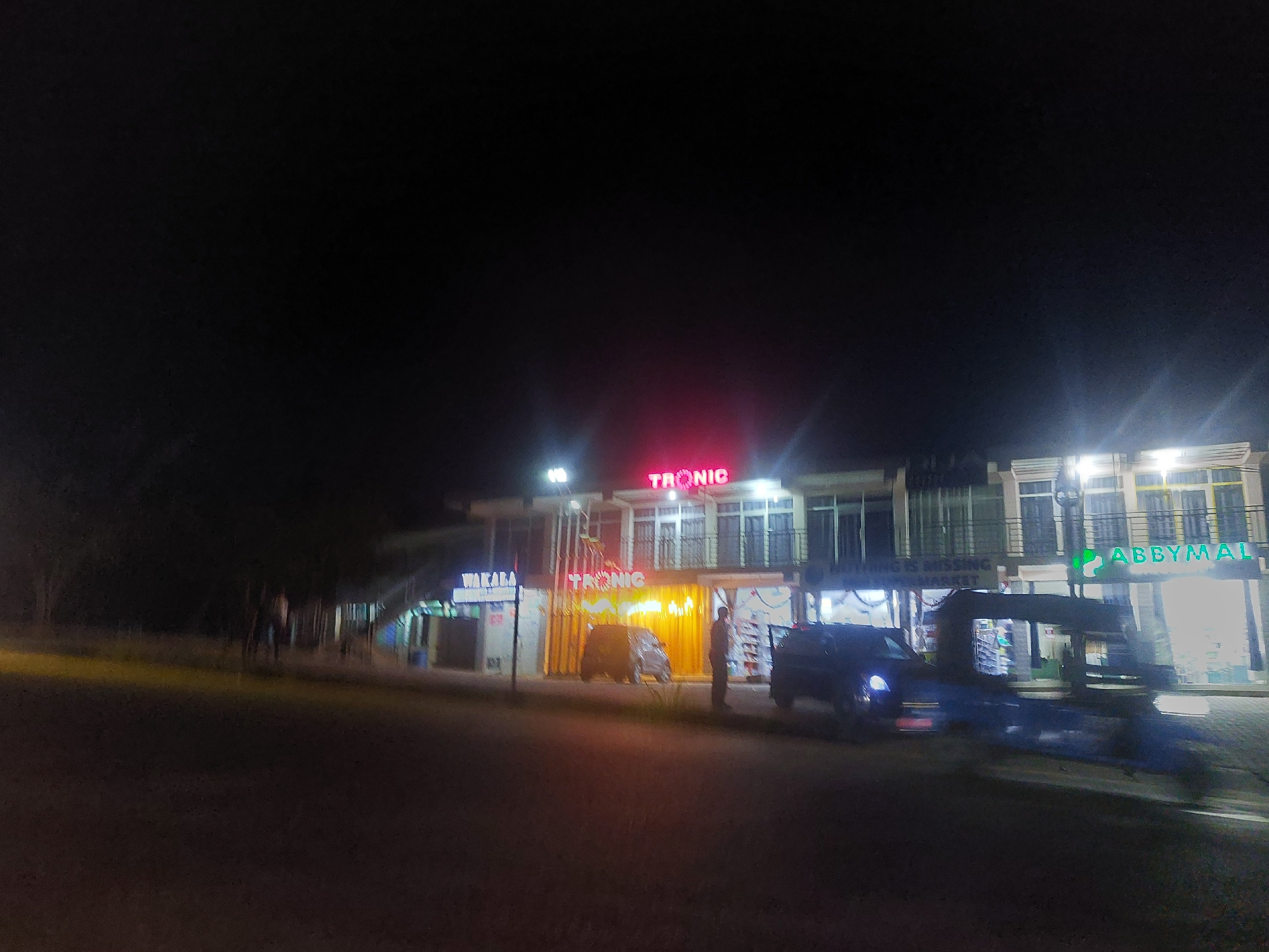 At night in Dar es-Salaam, Tanzania, a multi-story commercial building is visible. The building has signage for "TRONIC" in large, red neon letters on the upper level and a smaller sign above the entrance. Next to "TRONIC" is a sign that says "ABBYMAL." A person stands in front of the storefront and there are two automobiles in front of the building. The ground in the foreground appears to be a road surface. The overall lighting is dim.