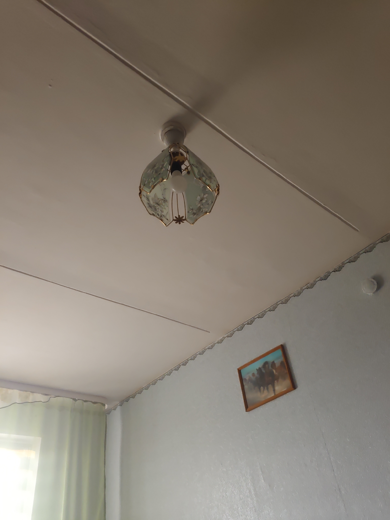 The image shows an interior view of a room, possibly a residential apartment in Курашим, Russia. The ceiling is white with a visible seam running diagonally across it. A light fixture with a decorative glass shade, featuring floral patterns in light green and gold trim, is attached to the ceiling. Inside the fixture, a light bulb is visible. On the wall below the ceiling, a framed picture depicting several horses in motion is hung. The wall has a textured wallpaper in a light gray color. A scalloped decorative border is visible where the ceiling meets the wall. To the left, a window is partially visible, covered by light green curtains. The overall impression is that of a simple, lived-in interior space.