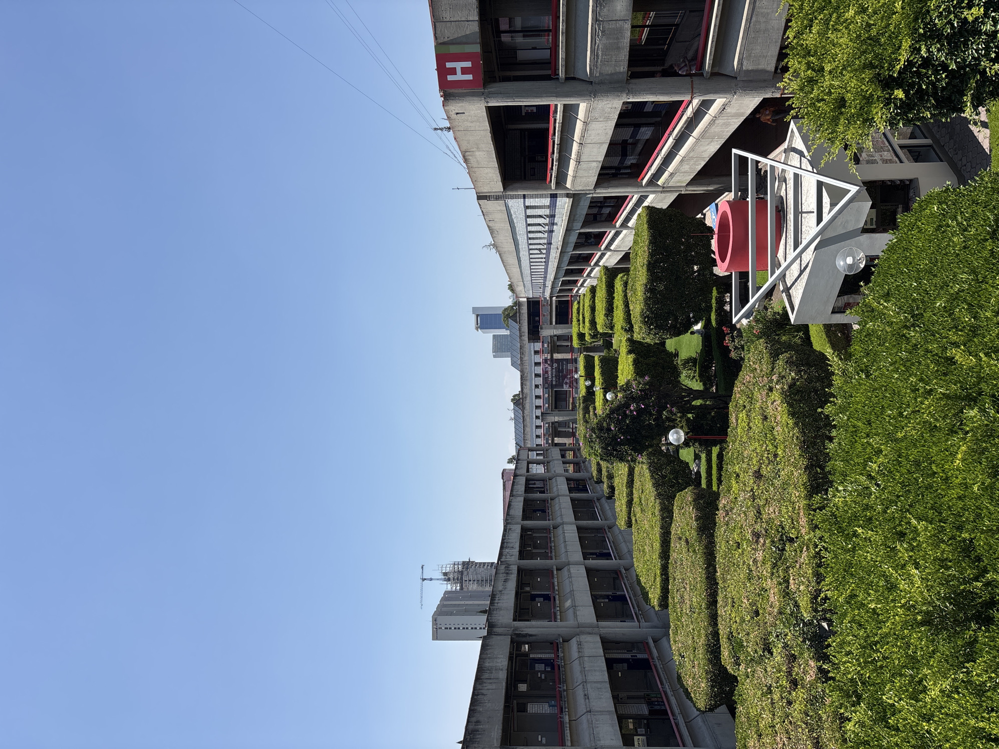 The image depicts the architectural landscape of the Universidad de las Américas Puebla (UDLAP) in Puebla, Mexico. The view showcases modernist-style buildings with concrete structures and horizontal lines. There are rows of manicured, rectangular shrubbery which add green contrast to the gray concrete. A red, cylindrical art installation is visible within a white, triangular frame. The buildings have the letter "H" on them. There is a partial view of the sky which is a clear blue. There is evidence of other buildings beyond the UDLAP campus, including a building under construction.