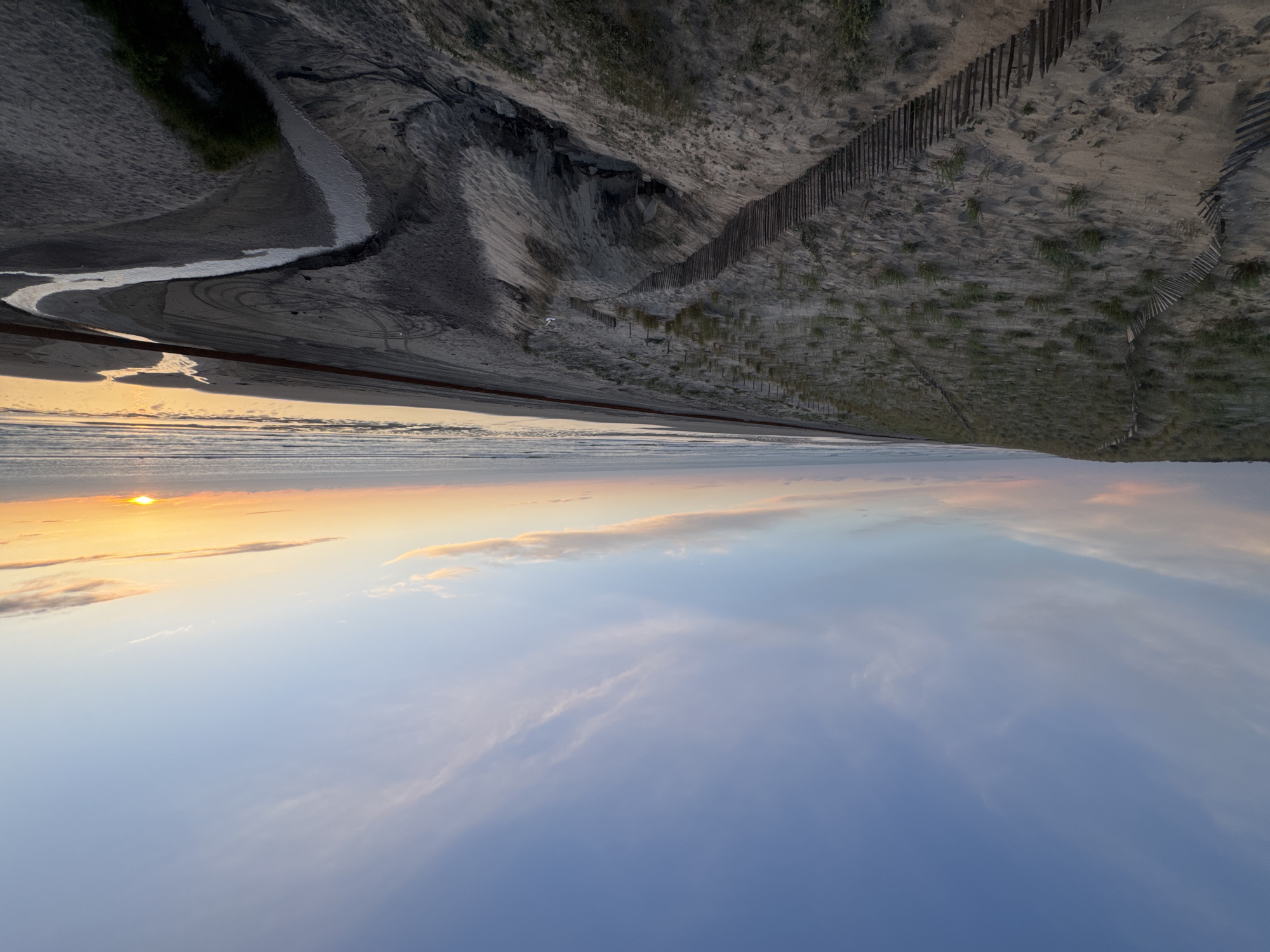 This image portrays a beach scene at Soulac-sur-Mer, France, during either sunrise or sunset. The top half of the image shows a landscape of sand dunes reinforced by wooden fencing. A small stream or waterway snakes its way through the sand towards the sea. Tire tracks mark the sandy surface. The bottom half depicts the sky and the ocean. A yellow sun is visible near the horizon, casting a golden hue on the water and the clouds. The sky transitions from a pale yellow-orange near the horizon to a light blue in the upper part of the image, with wispy clouds scattered throughout. The horizon line separates the water and the sky. The image is oriented in an unusual vertical manner, which emphasizes the height and the scale of the dunes in relation to the open sky and sea.