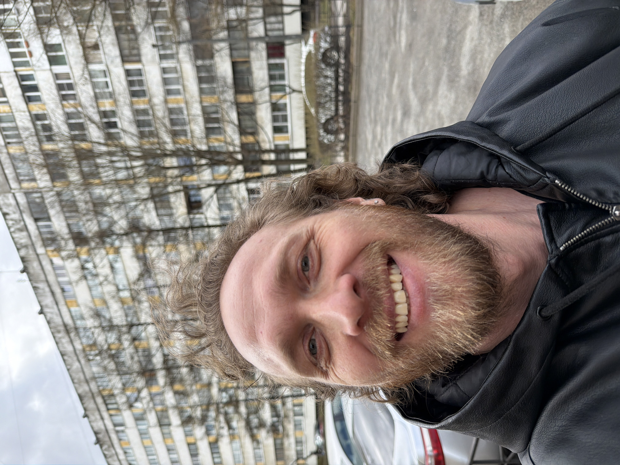 A Caucasian man with light-brown curly hair and a thick beard is the primary subject of the image. He is wearing a black leather jacket with the zipper partially open, suggesting cool weather. His face is turned towards the camera, and he is smiling, revealing his teeth.
Behind the man, a large, multi-story building with a repetitive pattern of windows is visible. The building's architectural style appears to be typical of Soviet-era construction, possibly in a residential area. The presence of bare trees suggests it might be late autumn or early spring.
The image was taken in the city of Saint Petersburg, Russia.