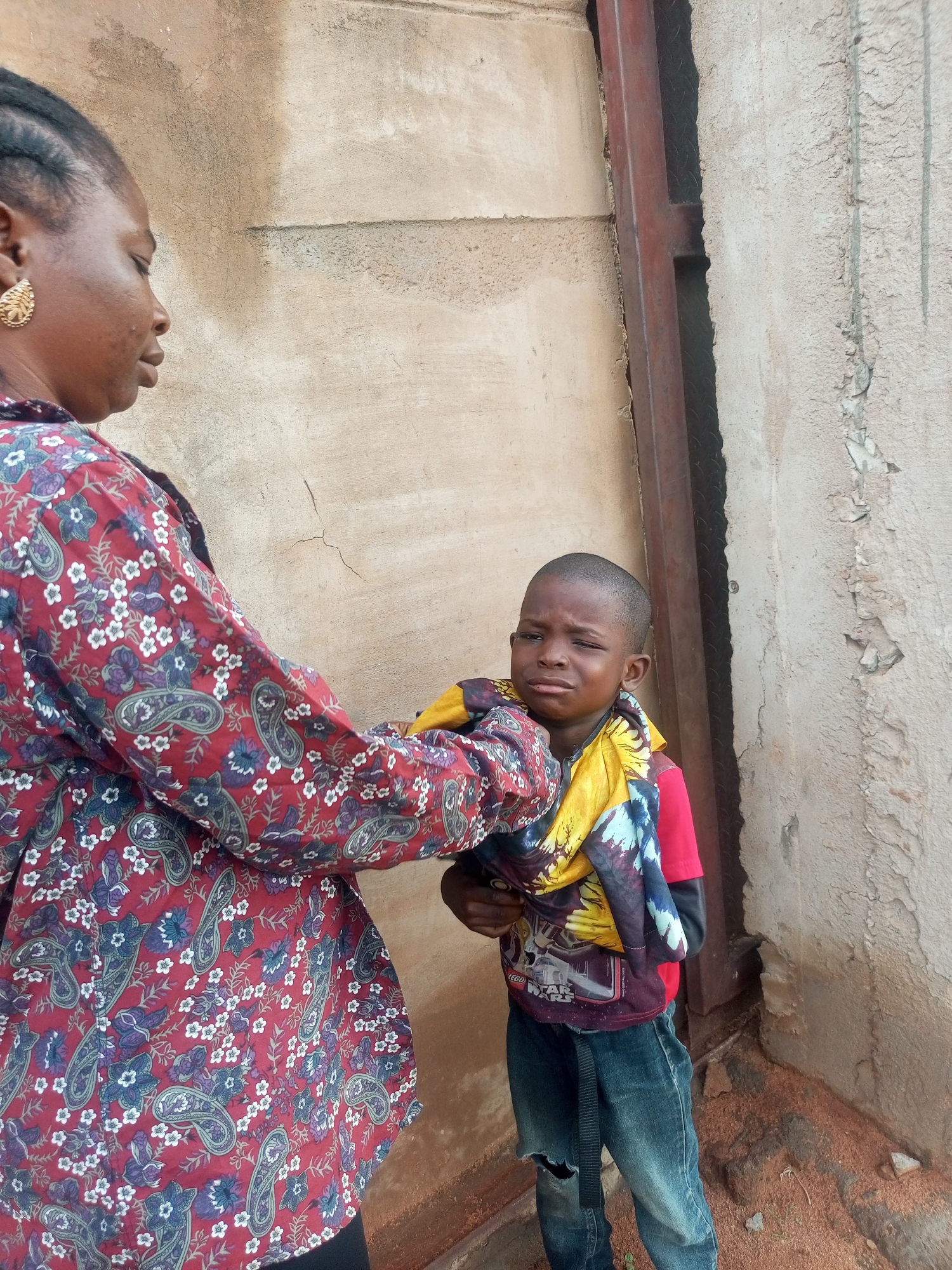 An adult woman in a patterned red dress with a floral and paisley design and a gold earring is holding a young boy. The boy is wearing a red shirt, blue jeans with a ripped knee, and a colorful scarf around his neck. He appears to be crying. They are standing outdoors in front of a textured concrete wall, with a vertical metal beam to the right of them. The ground is covered in reddish sand and small stones. The scene is consistent with an outdoor location in a residential or unpaved area of Tempure, Nigeria.