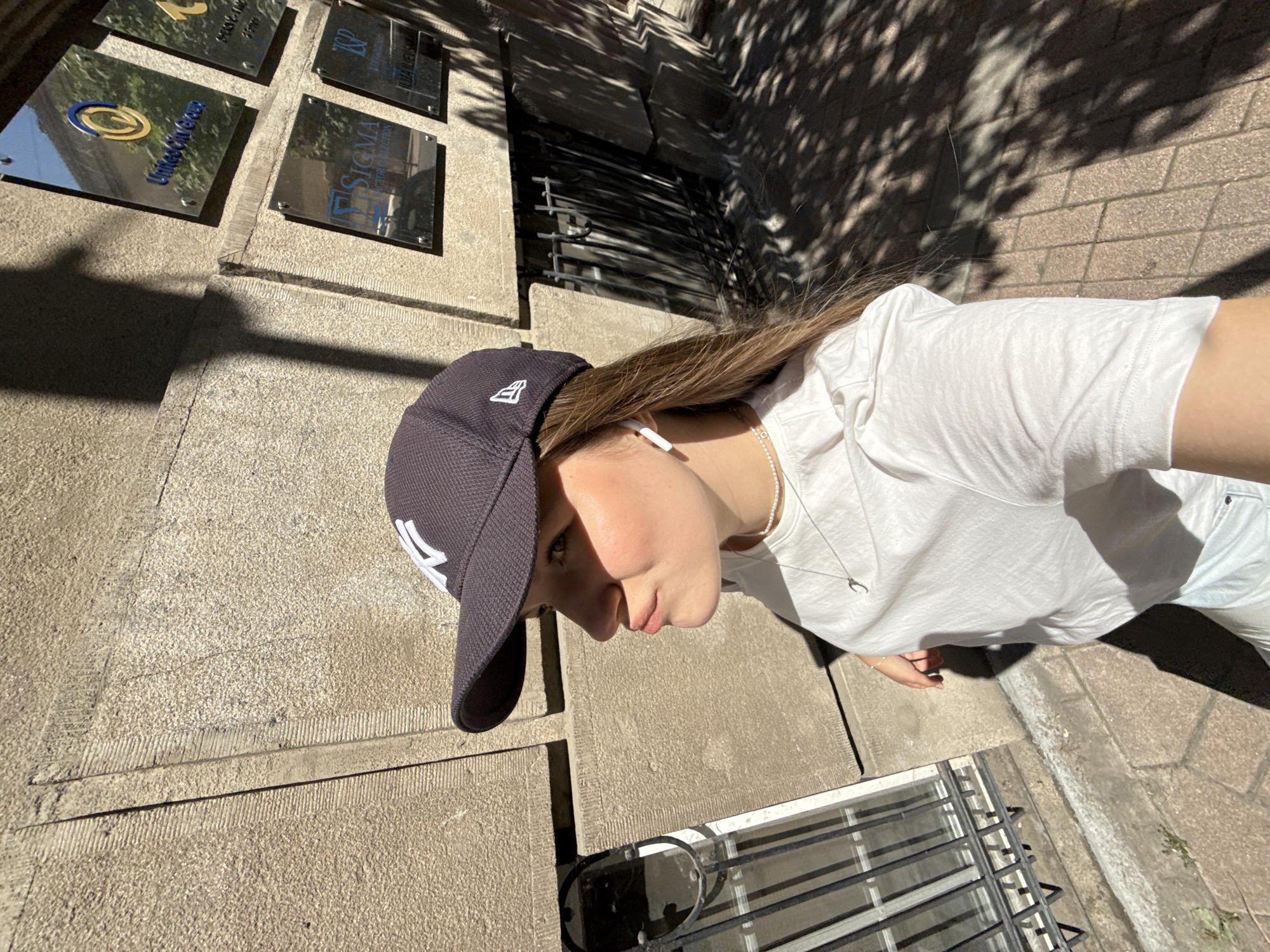 A young woman wearing a New York Yankees baseball cap and a white T-shirt stands on a sidewalk in Belgrade, Serbia. She is positioned in front of a building with multiple metallic nameplates, including "United Oil Group" and "Sigma Group Selection." Sunlight casts shadows from trees onto the sidewalk and building facade. A barred window is visible near the woman's feet.