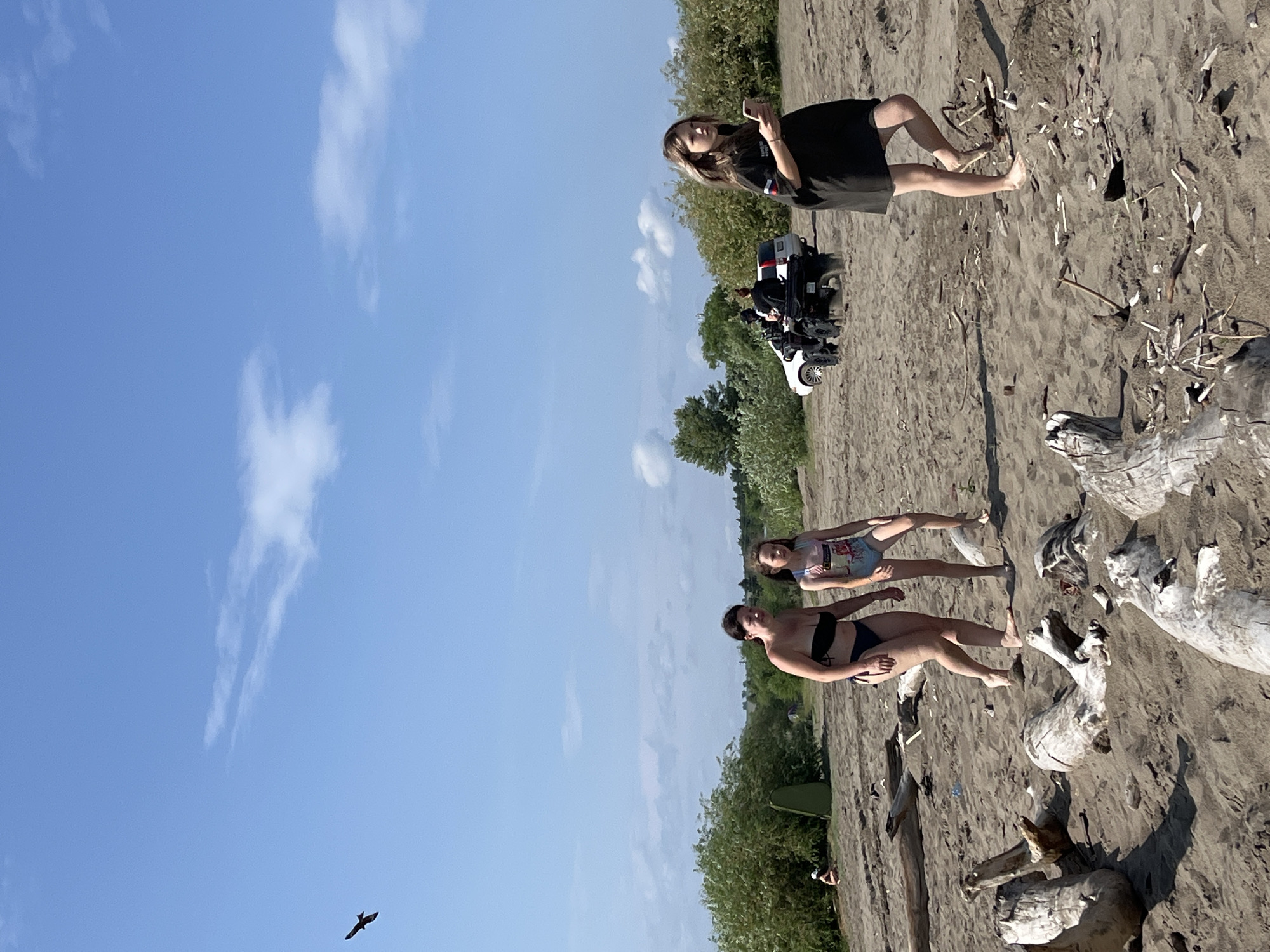 The image depicts a sunny day at a sandy beach in Krasnoturansk, Russia. The sky is a vibrant blue with scattered white clouds. In the foreground, a person wearing a black t-shirt and shorts is standing, holding a mobile phone. Behind them, a white all-terrain vehicle (ATV) is parked on the sand. Further back, two people are lying on the beach, one in a bikini and the other in a black swimsuit. The beach is characterized by sand and scattered driftwood. Lush green trees line the background, suggesting a natural, outdoor setting. The overall atmosphere appears relaxed and casual, indicative of a leisurely day at the beach. The time of day seems to be late afternoon, given the quality of light.