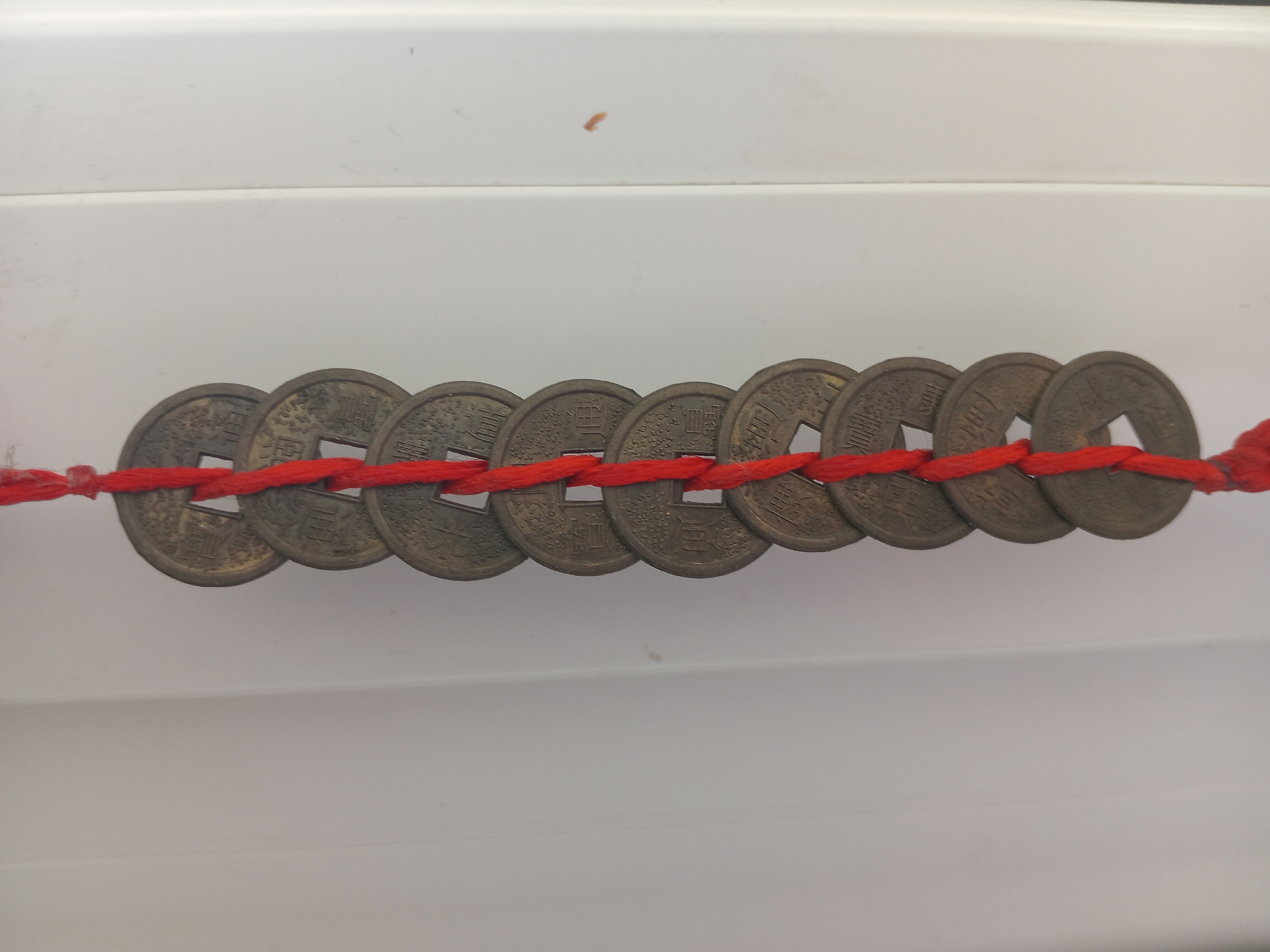 A close-up, vertical shot shows a string of ten antique Chinese coins, tied together with a red cord. The coins are circular with a square hole in the center and have Chinese characters inscribed on them. They are arranged in a linear fashion, hanging vertically. The background is a plain, light-colored surface, likely a wall, with a subtle vertical seam running down the right side of the image. The lighting is even, highlighting the texture and details of the coins. No people or further environmental context are visible.