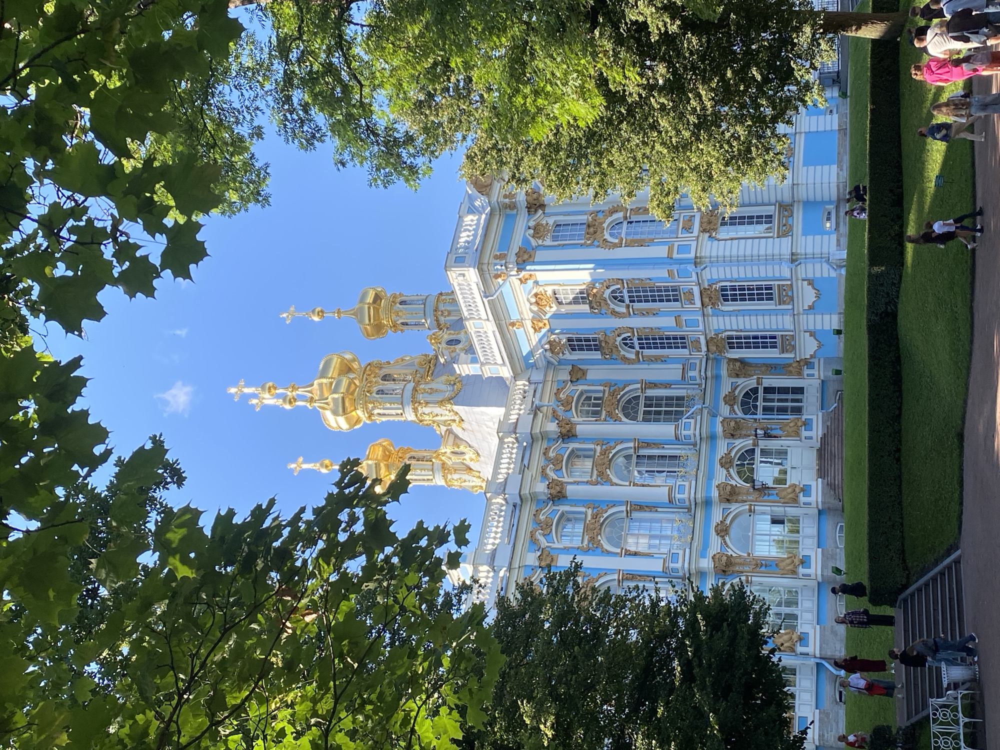 The image depicts the Catherine Palace, located in the city of Pushkin, Russia, on a clear, sunny day. The palace is a grand Baroque structure with a striking white and light blue facade adorned with gold detailing. Multiple golden domes rise from the roof, topped with crosses. The palace is framed by lush green trees, with their leaves partially obscuring the blue sky. In the foreground, several people are visible walking on pathways and on a grassy lawn. Some are ascending or descending stone steps leading to the palace entrance. The scene suggests a tourist or visitor setting, with individuals casually dressed and engaging in recreational activities in the palace grounds. The overall atmosphere is one of historical grandeur and natural beauty.