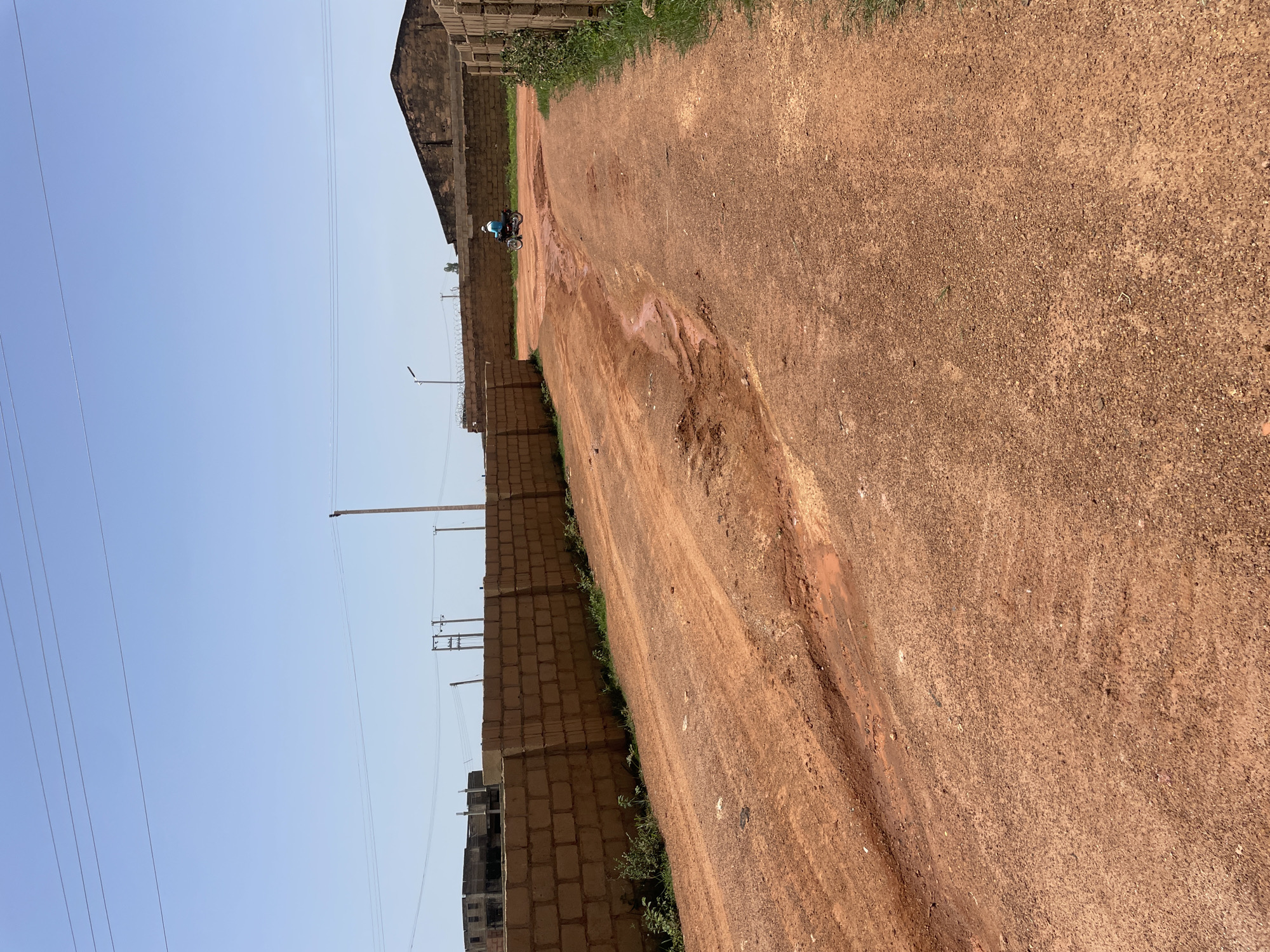A dirt road in Bar Jirgi Summa, Nigeria, with a red clay surface showing ruts and a small muddy stream. A person is riding a motorcycle down the road, and there are brick walls lining the sides. In the background, there are buildings and power lines against a clear blue sky.