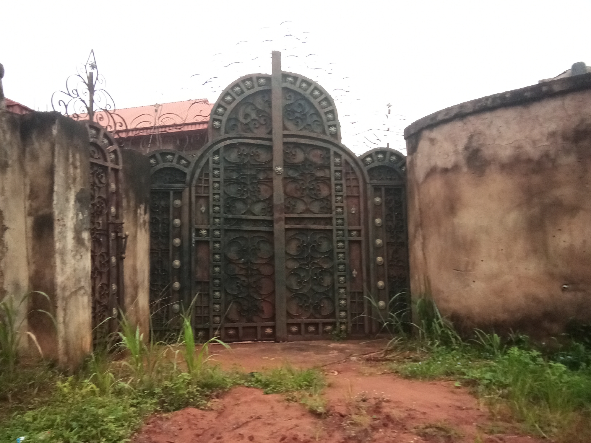 The image shows an ornate, dark metal gate in front of a building with a red tile roof. The gate is tall and features intricate scrollwork and geometric patterns. It is flanked by rough concrete walls, one on the left and another on the right which appears to be a part of a building. The ground in front of the gate is red earth and covered with sparse green vegetation and tall grass. The sky is overcast and a pale grey. The scene is likely a residential property in Oko Town, Nigeria, given the architectural style and the surrounding environment. There are no people or animals visible in the image, and no apparent actions or interactions are taking place.