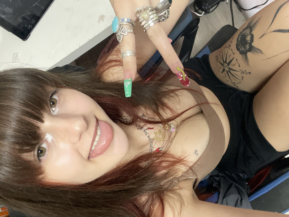 A young woman with brown hair and bangs, fair skin, and green eyes looks at the camera with her tongue slightly out. She wears several rings on her fingers, a necklace with various charms, and a brown top with black shorts. Her fingernails are decorated, one with a cocktail glass and the other with cherries. A tattoo of a flower and a compass adorns her thigh. A white substance appears on her nose. The background is a mix of a light-colored surface and a black chair.