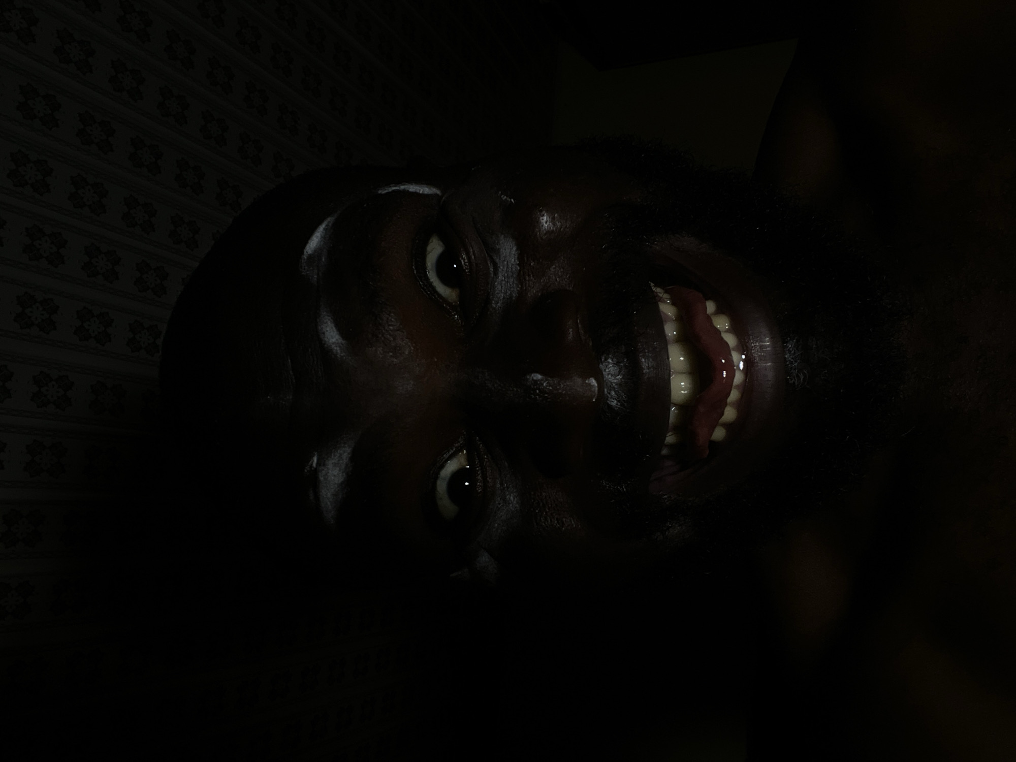 A close-up, slightly angled, low-light photograph shows the face of a person with dark skin. The person's mouth is open, revealing teeth and tongue. The background consists of a patterned surface. The location is unknown.