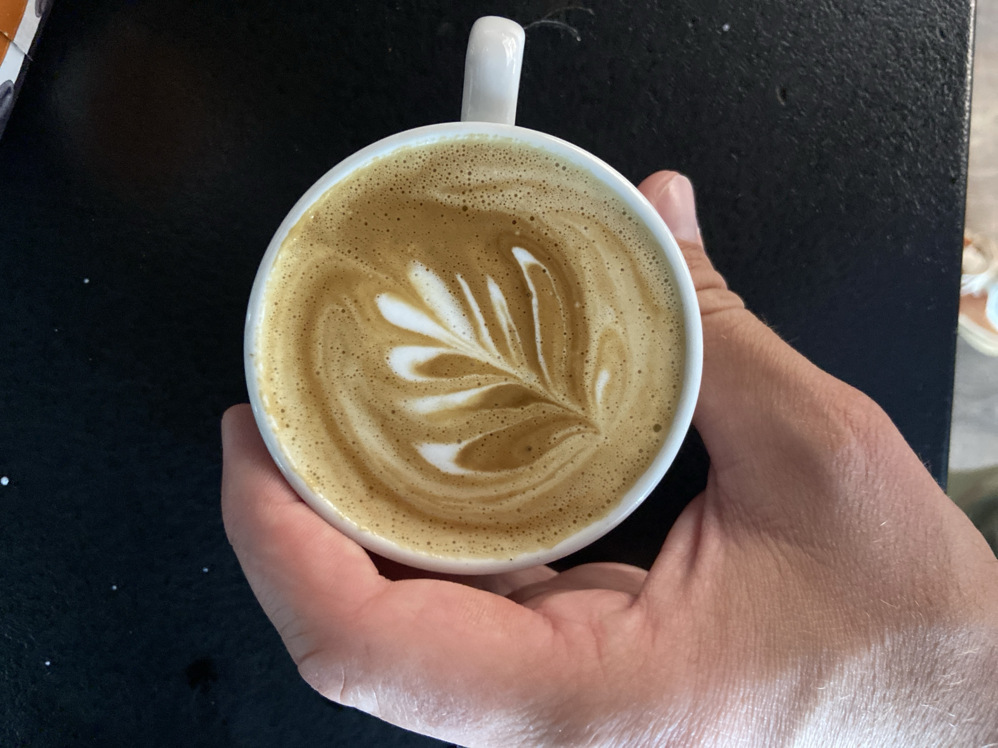 A person is holding a white coffee cup with latte art in the shape of a leaf. The background is dark and blurred, with a partial view of a table surface. The location, Petrovac centar, Montenegro, cannot be verified from the image.