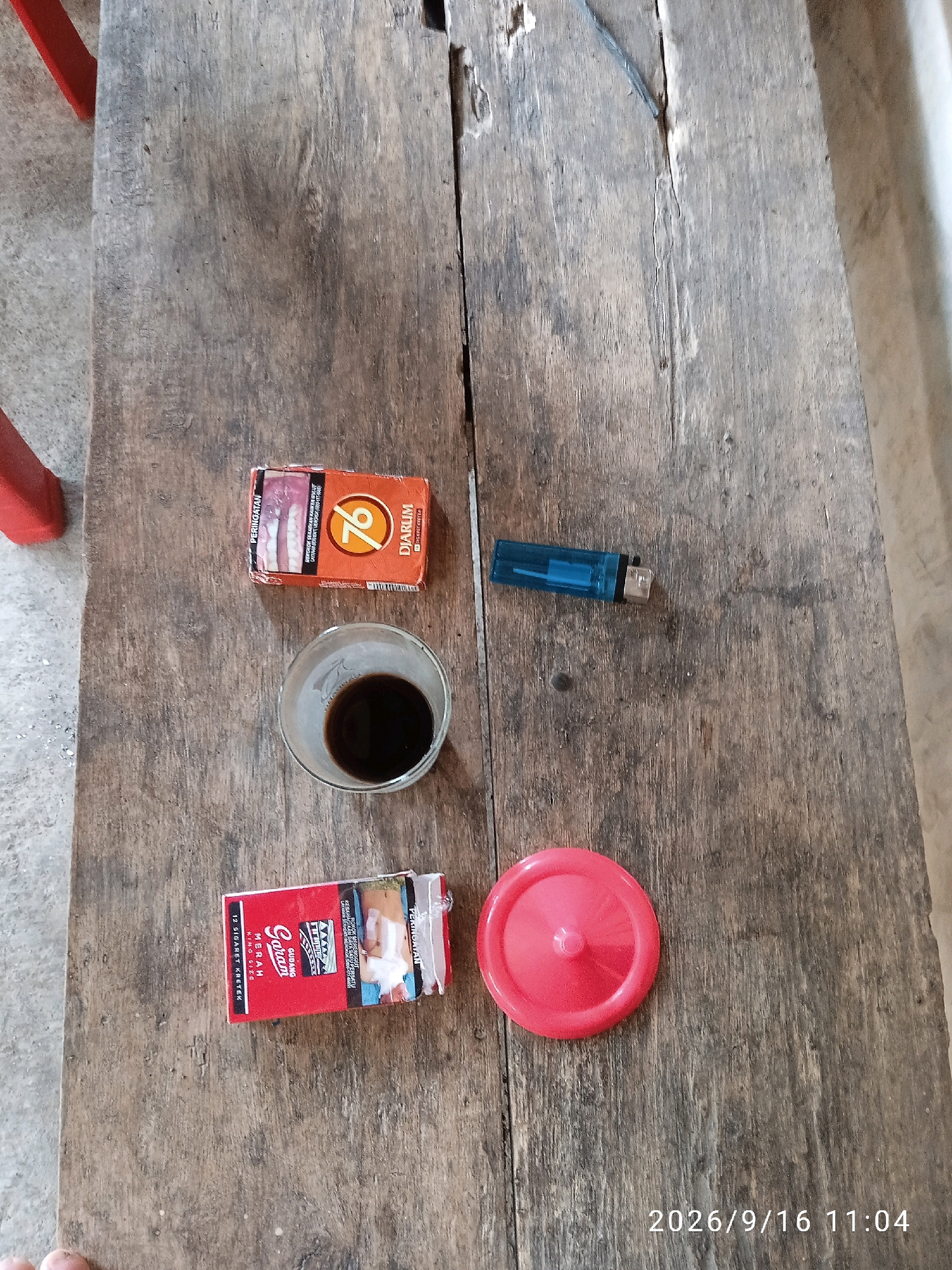 A top-down, close-up shot shows a wooden table with several items on it. In the upper left quadrant is a red-and-orange cigarette pack labeled "76 DJARUM PREMIUM" with a warning label on the left side. To the right of the cigarette pack is a clear glass containing a dark liquid, likely coffee. Below the cigarette pack, a red cigarette pack labeled "Gudang Garam MERAH" with the text "KING SIZE" is visible. To the right of the glass, a blue disposable lighter rests on the table. In the lower right quadrant, a bright pink circular object, possibly a lid or cap, is placed on the table. The wooden table surface is worn and appears to be outdoors or in an open space, given the concrete-like material and red furniture legs visible in the background on the left. The timestamp "2026/9/16 11:04" is superimposed on the bottom right of the image. This scene is consistent with a casual setting in Kelurahan, Indonesia, where such items are commonly found.