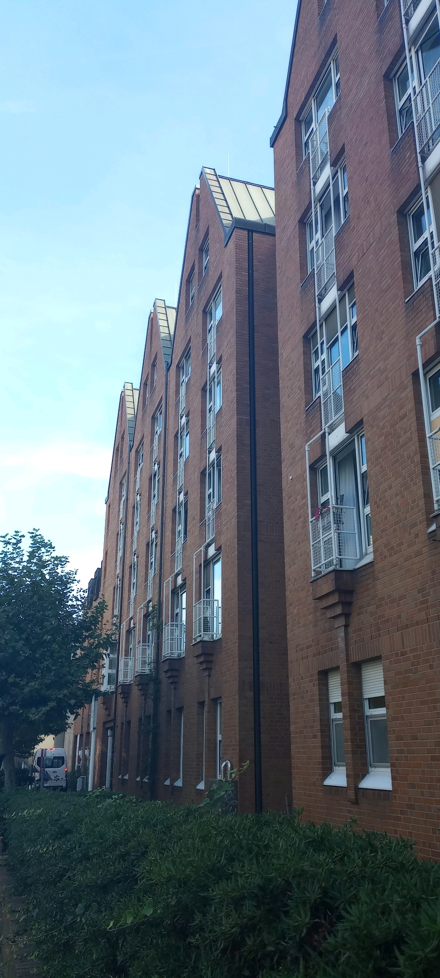 The image shows a residential building constructed of red brick, featuring a gabled roof with a metal finish. The building has multiple floors with numerous windows, some of which are equipped with small balconies with white metal railings. The windows are framed in white, and some have white roller shutters. The architecture appears modern, with a stepped effect at the base of the balconies.
To the left of the building, a green tree with dense foliage is visible. In the background, a white van with tinted windows is parked near the building. A small patch of sidewalk and a street are also partially visible. The ground in front of the building is covered with a thick hedge of dark green bushes. The sky above is clear and light blue, indicating fair weather.
This building is located in the Scharnhorst district of Dortmund, Germany, specifically on the street "In der Kolvenburg". The image captures the exterior of a residential complex, highlighting the architectural style and its surrounding urban environment.