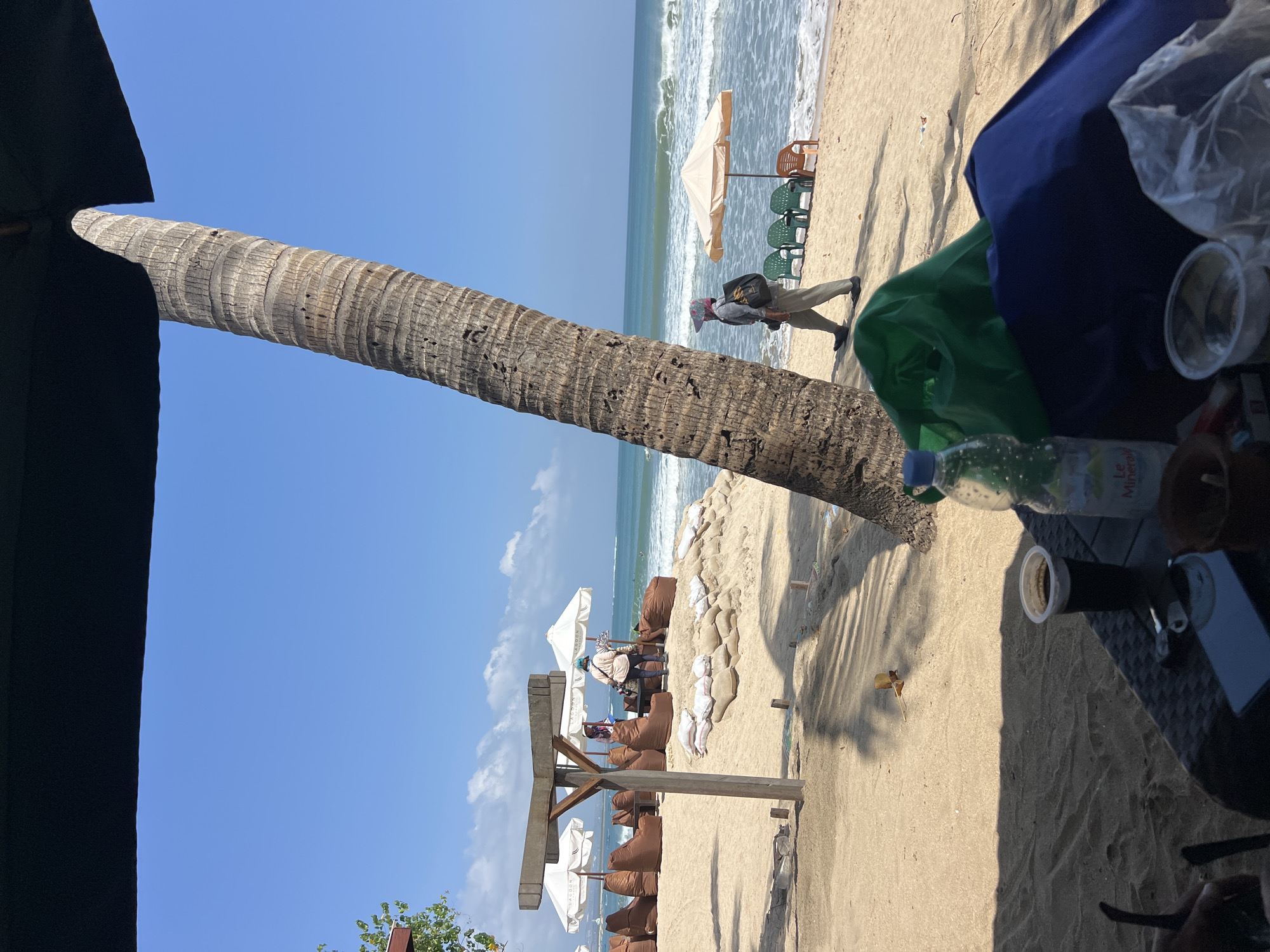 The image depicts a sunny day on a beach in Kuta, Indonesia. In the foreground, a table is set with a drink in a plastic cup, a bottle of water, and various other items. A large palm tree trunk dominates the mid-ground, leaning towards the right. Behind the palm tree, the beach is lined with brown beanbag chairs and white umbrellas. Two individuals are visible near the seating area, one standing and one seated. Further back, the ocean waves crash onto the shore under a clear blue sky. In the distance, more umbrellas and chairs are visible on the beach, and a person is walking along the shoreline. The scene suggests a relaxed atmosphere at a beachside establishment.