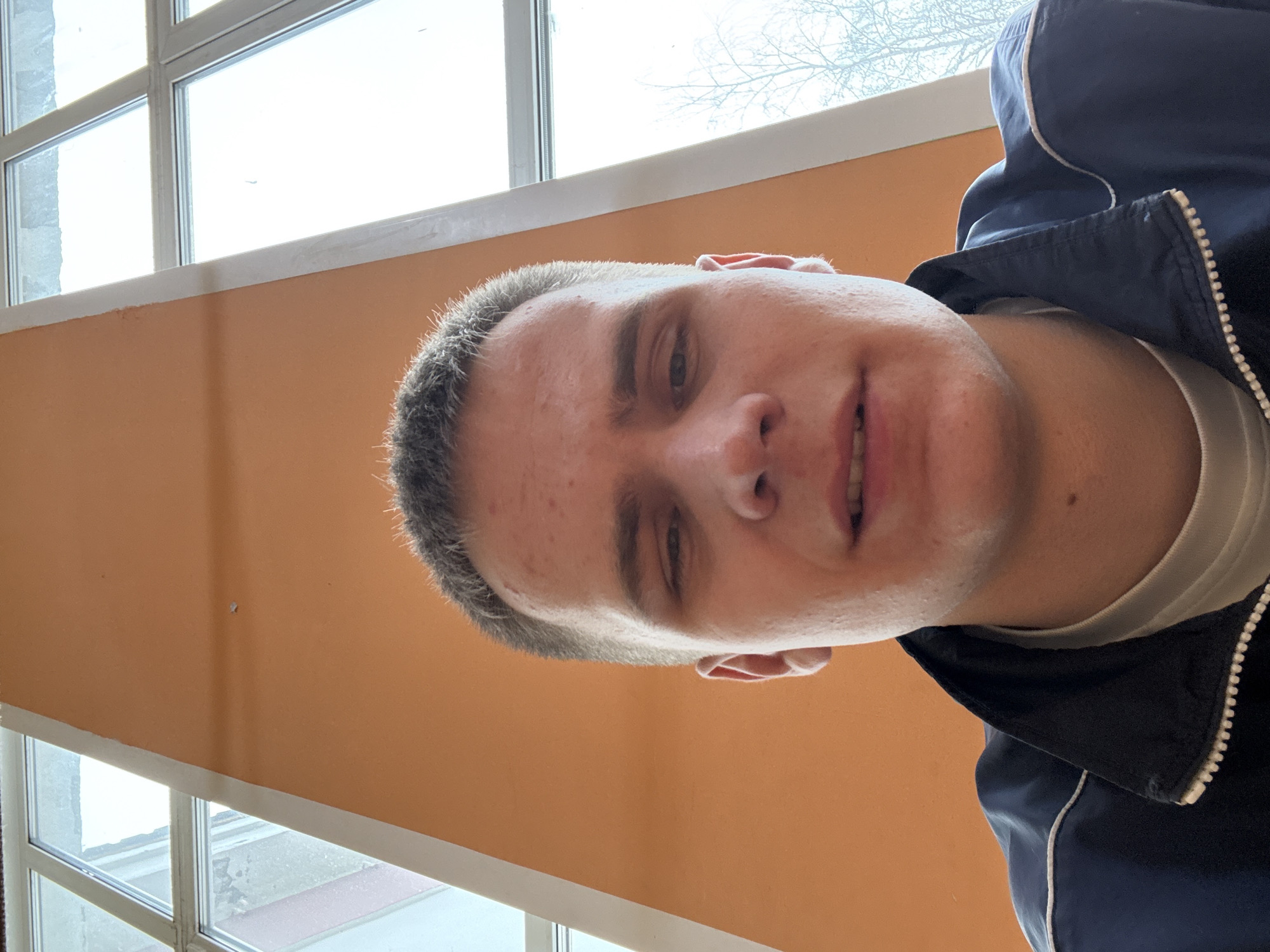 A man with short, graying hair is centered in the image. He wears a white t-shirt under a dark blue jacket with a white stripe. He is indoors with an orange wall behind him. A window with a white frame is visible on the left side, showing a view outside with bare tree branches. The image was taken at an angle. There is no way to determine the location in the city of Chaikovsky, Russia.