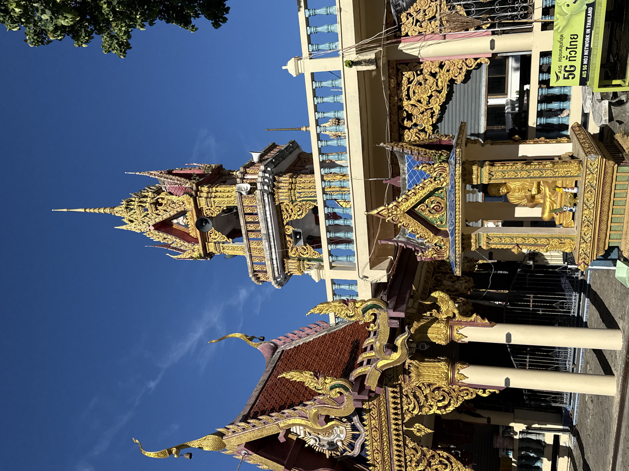 The image depicts the exterior of a Buddhist temple in Baan Plai Laem, Thailand, under a clear blue sky. In the foreground, a golden altar with a seated Buddha statue is prominently displayed. The temple itself features ornate golden decorations on its roofs, pillars, and walls, characteristic of Thai temple architecture. A tall bell tower with a golden spire rises in the background, adorned with decorative elements and equipped with loudspeakers. The temple complex also includes a balcony with a blue railing. In the lower right corner of the image, a lime green sign advertising "AIS 5G" with a smiling cartoon character is visible. Lush green trees are seen to the right and partially framing the top of the image. The ground is paved with stone tiles. There are no people visible in the image.