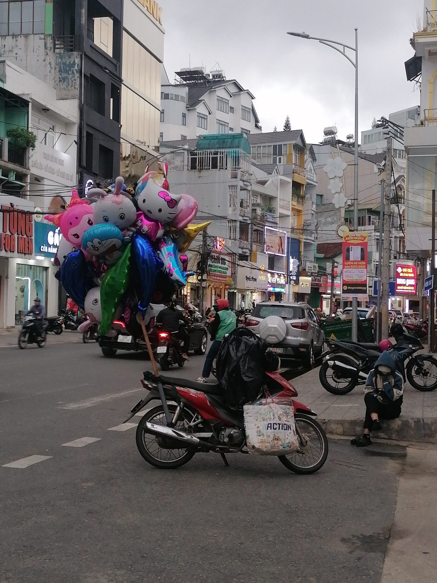 The image depicts a street scene in Da Lat, Vietnam. The weather is overcast. In the foreground, a red motorcycle with a large "ACTION" bag is parked on the road. A person wearing a hooded jacket is sitting on the curb nearby, appearing to look at their phone.
In the middle ground, a motorbike carries a large bundle of balloons in various cartoon shapes. Behind this vehicle, there is a silver SUV and other motorcycles on the road.
In the background, a street with multiple buildings and businesses is visible, identifiable by colorful signage. A street light is visible to the right. Power lines crisscross overhead. Banners and advertisements are displayed on buildings and poles along the street, and there are several people and motorcycles on the street.