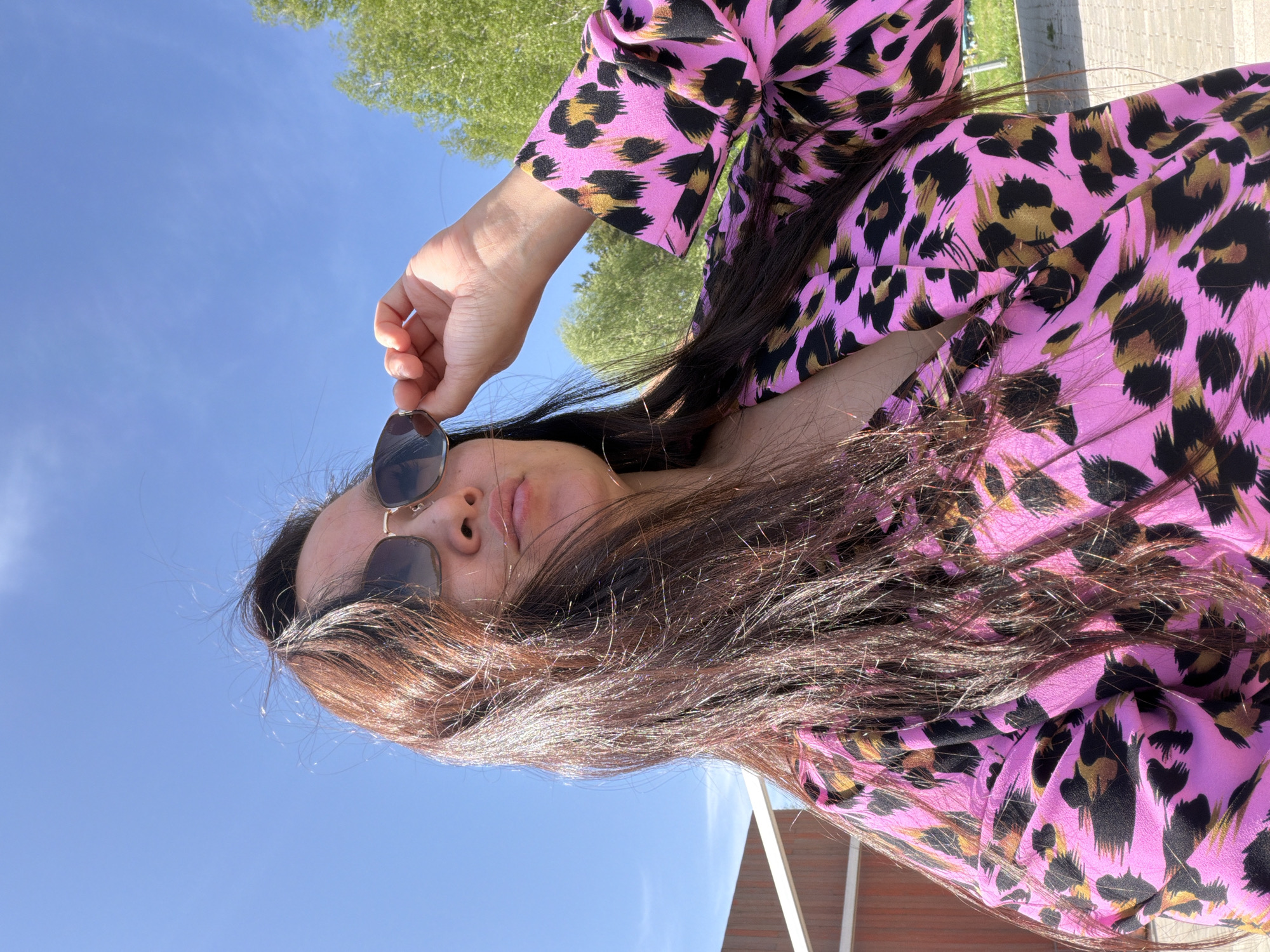 A woman wearing a pink and black leopard print top is holding sunglasses in her right hand, positioned near her face. She is outside with a blue sky overhead and green trees visible in the background. Parts of a building or structure are visible in the lower part of the image.