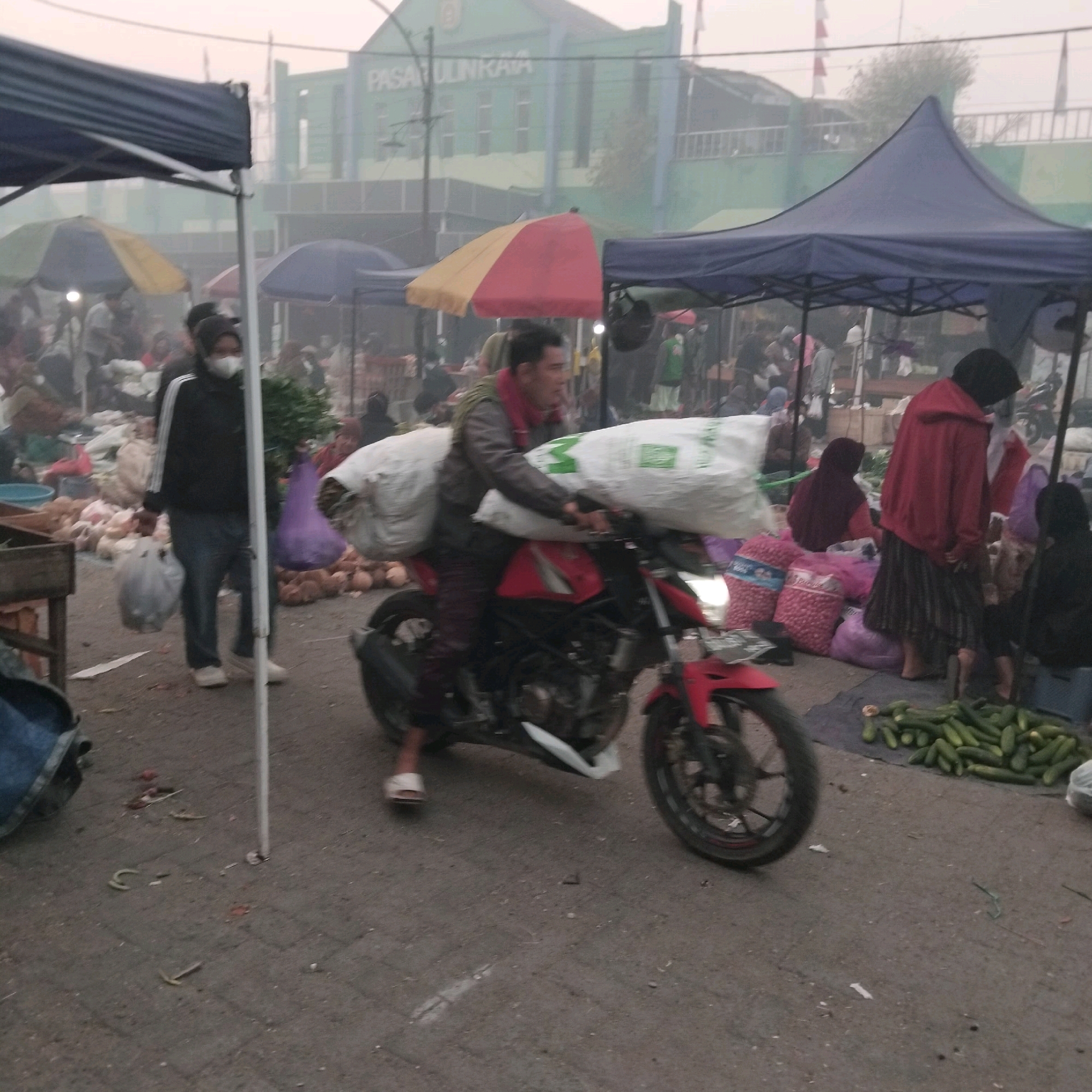 The image depicts an outdoor market scene in Banjarbaru, Indonesia, with a focus on a man riding a red motorcycle loaded with two large, white sacks. The man is wearing a dark jacket with a red scarf and dark pants, and he is looking to his right. The motorcycle has its headlights on. In the foreground, to the right of the motorcycle, is a pile of cucumbers. To the left of the motorcycle, a person wearing a dark jacket and jeans walks away from the camera, carrying two plastic bags. A large blue tent is visible in the upper left corner, and a yellow and red umbrella is also present. In the background, a green building with the text "PASAR ULINKAIA" is visible. The market appears to be busy, with other people and stalls blurred in the background, suggesting a hazy or dusty atmosphere. The ground is paved with asphalt.