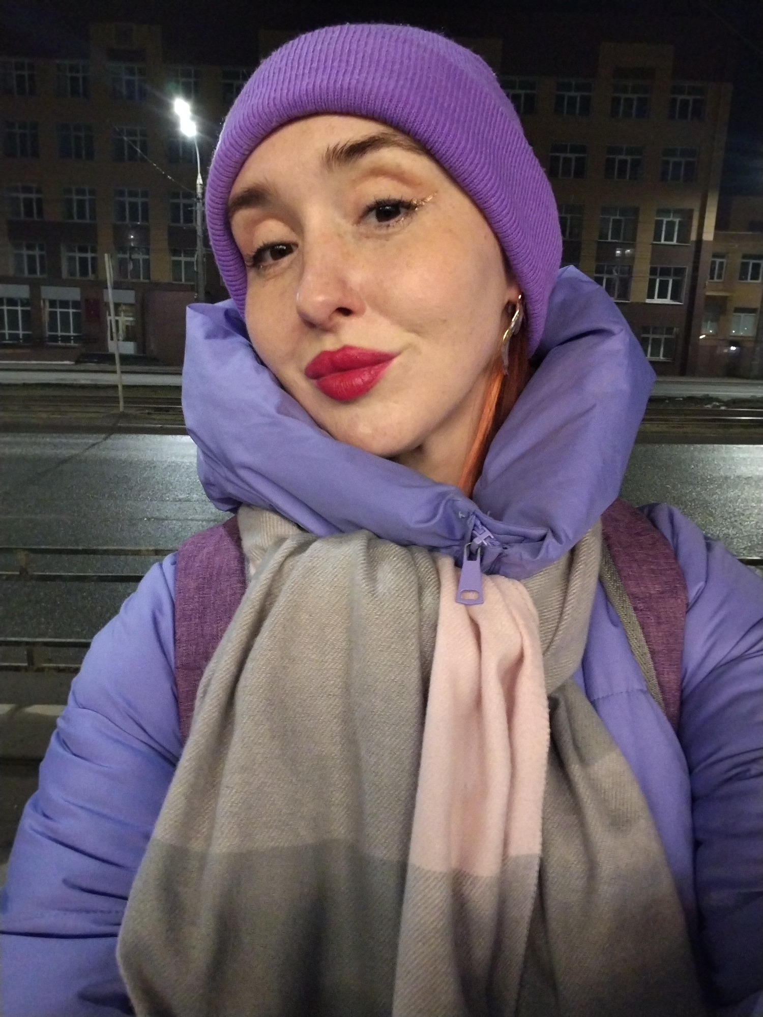 A woman is taking a selfie outdoors at night. She is wearing a purple beanie, a purple puffer jacket, and a grey and pink scarf. She has red lipstick and visible earrings. Behind her is a city scene with buildings, a street lamp, and a street. Based on the buildings and streetscape, the location is consistent with Izhevsk, Russia. There are railroad tracks and a low railing.