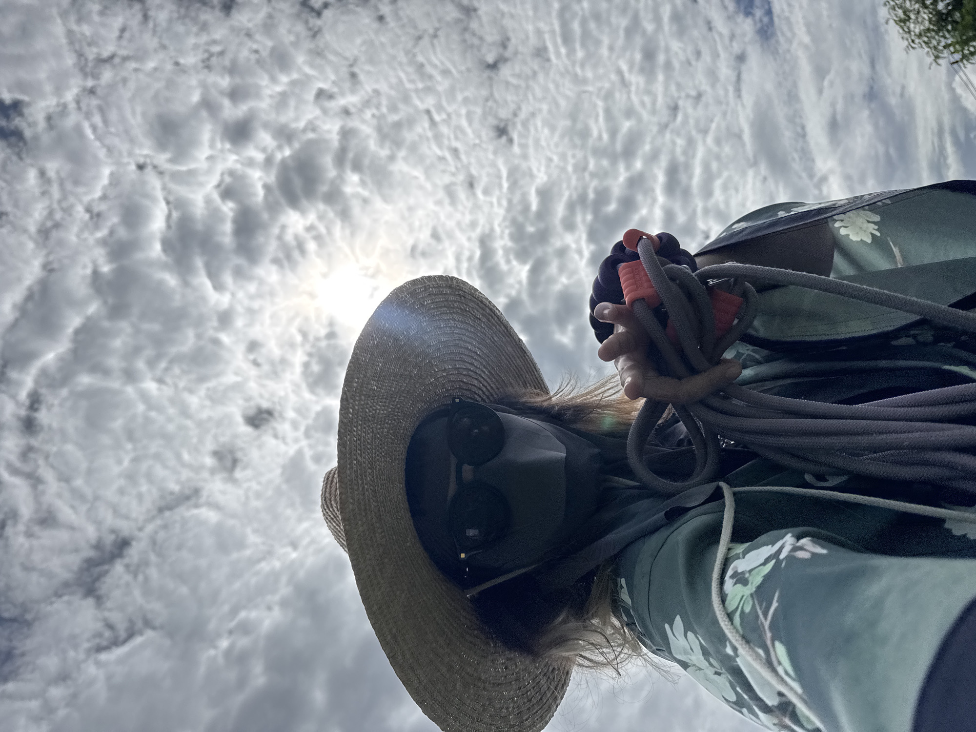 A person wearing a wide-brimmed straw hat, sunglasses, and a dark face mask is holding a coiled rope in their right hand. They are wearing a green, floral-patterned garment. The background is a cloudy sky with the sun partially obscured by clouds, creating a lens flare effect. The low-angle shot suggests the photo was taken from below the subject, looking up towards the sky. The location is stated to be in the city of Karon, Thailand.