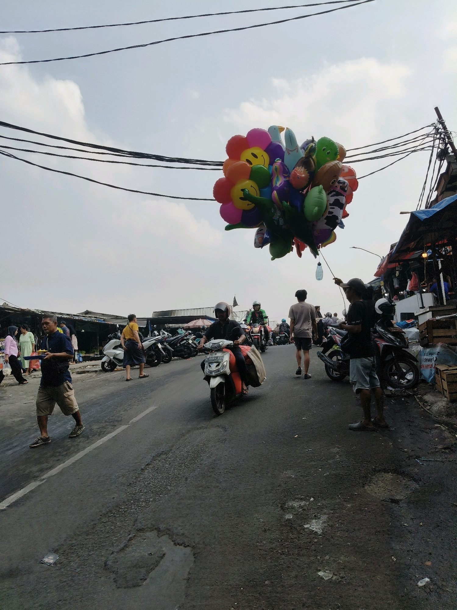 This is a street-level, eye-level photograph taken on a paved road in South Tangerang, Indonesia, under an overcast sky. The scene depicts a bustling market or roadside environment. Dominating the upper half of the frame is a cluster of colorful, cartoon-shaped balloons, including smiley faces, a cow print, and animal shapes, held aloft by a thin string. Several power lines are strung across the sky.
On the road, a man on a white scooter is riding towards the viewer, wearing a helmet. To his left, a man in a dark blue shirt and khaki shorts walks away from the camera, carrying a yellow object. Further left, other pedestrians and market stalls with awnings are visible. To the right of the road, several parked motorcycles are lined up, and a man in a dark t-shirt and light shorts stands holding a string attached to a bottle, potentially for the balloons. Beyond him, more market stalls and structures are apparent. The road surface is rough and shows signs of wear and tear, with several potholes. The overall impression is one of a lively, everyday scene in a busy Indonesian marketplace.