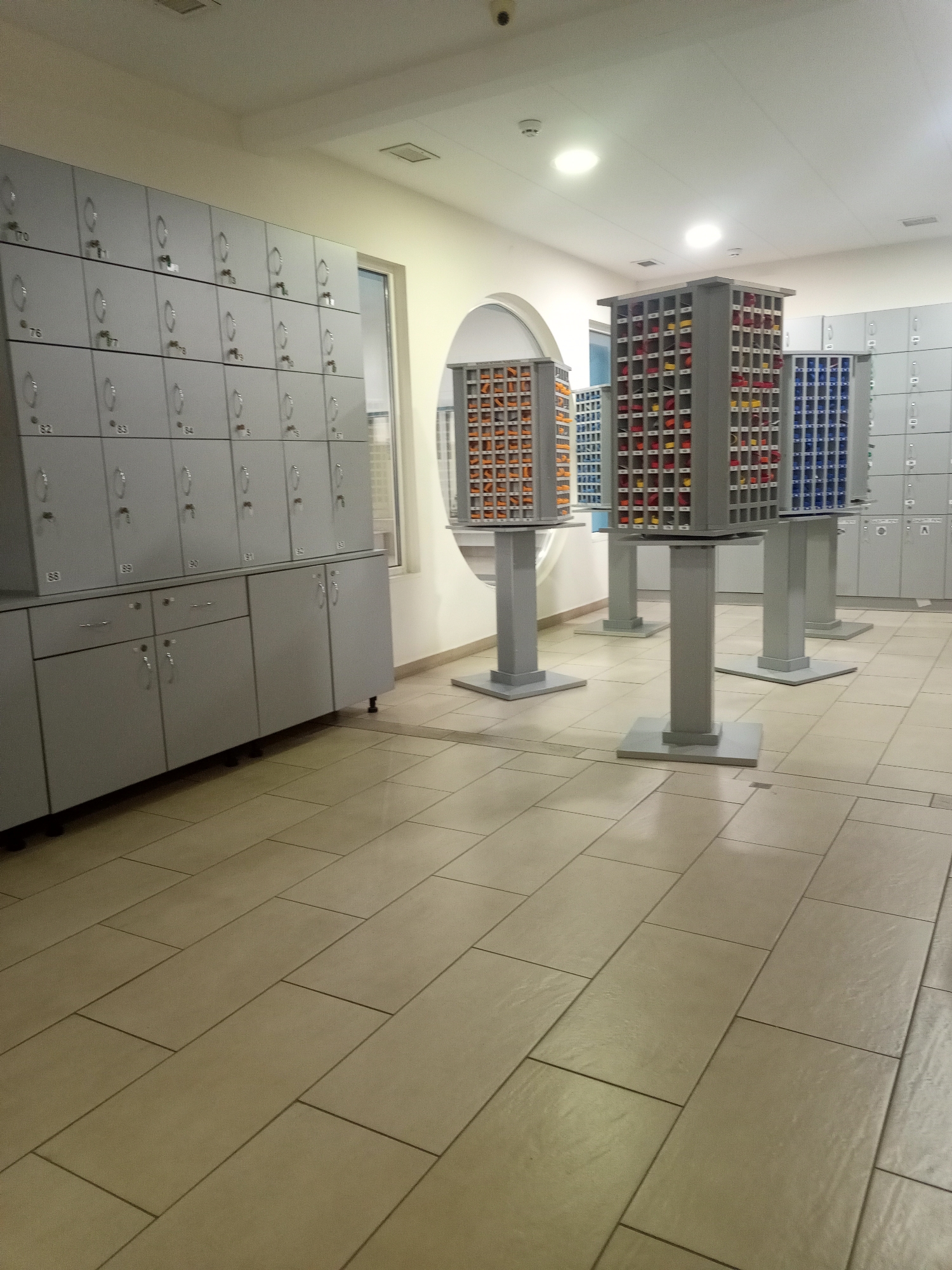 This media file displays an indoor scene featuring rows of grey metal lockers and several tiered storage units filled with colorful items. The storage units are square with multiple levels of compartments, some filled with orange, yellow, and blue items. These units are elevated on grey pedestals. On the left side of the image, a large bank of grey lockers is visible, with some doors open and others closed, each with a key. Below the lockers are several cabinets with drawers and doors. The floor is tiled with large, light-colored square tiles laid in a staggered pattern. The lighting suggests it is daytime, with overhead lights illuminating the space. There are no people visible in the image. The overall impression is of a locker room or storage area within a facility like a gym, pool, or a school. Some locker doors have visible numbers such as "70", "76", "82", "83", "84", "5", and "57", and "88", "89", "90", "91", "92". One of the storage units has text labels in red above each row of compartments.