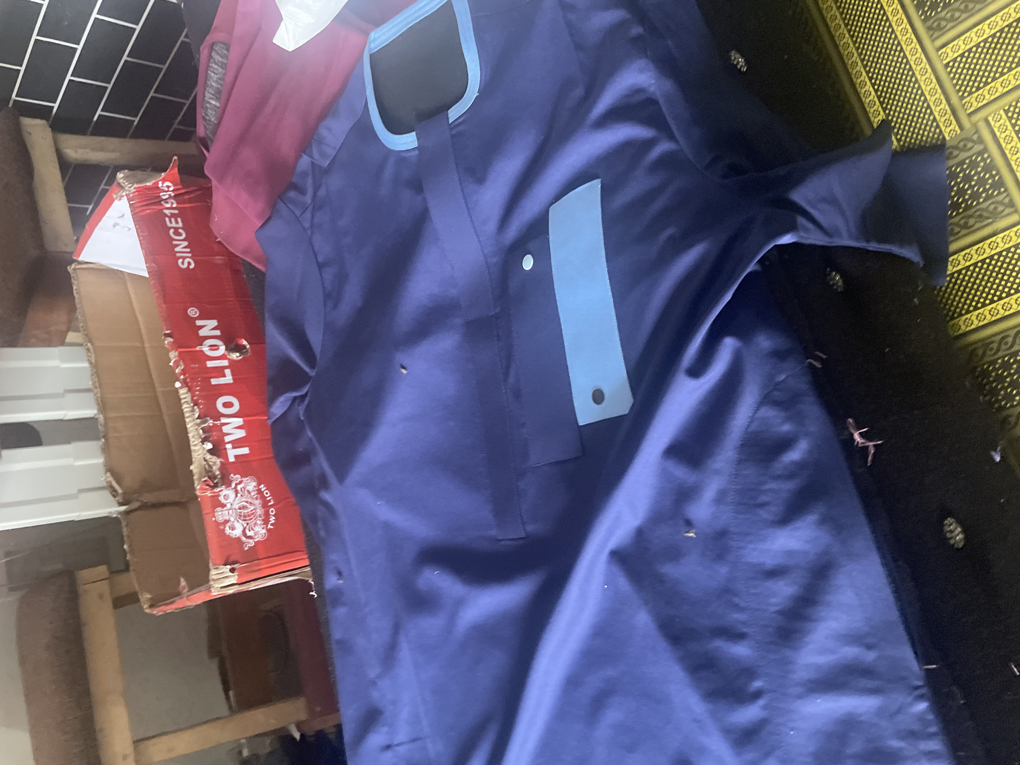 The image shows a close-up of a navy blue garment, possibly a kaftan or jubba, laid out on what appears to be a patterned rug. The garment has a contrasting light blue trim around the neckline and a light blue pocket on the chest, secured by two circular buttons. The fabric of the garment is smooth and appears to be made of a synthetic material, with some slight creasing. There are also a few small, dark specks on the fabric, possibly lint or dust.
In the background, to the left of the garment, a portion of a red cardboard box with white text is visible. The visible text reads "SINCE 1995" and "TWO LION" along with a logo that appears to be a coat of arms. Behind the box, a glimpse of a tiled wall with a black and white pattern can be seen. To the far left, part of a maroon-colored garment is also visible, draped over something unseen. The overall impression is of items being stored or displayed in a domestic or retail setting. There are no people visible in the image, and the lighting suggests an indoor environment, possibly during daylight hours. The context of Jalingo, Nigeria, provides a geographical setting for the scene.
