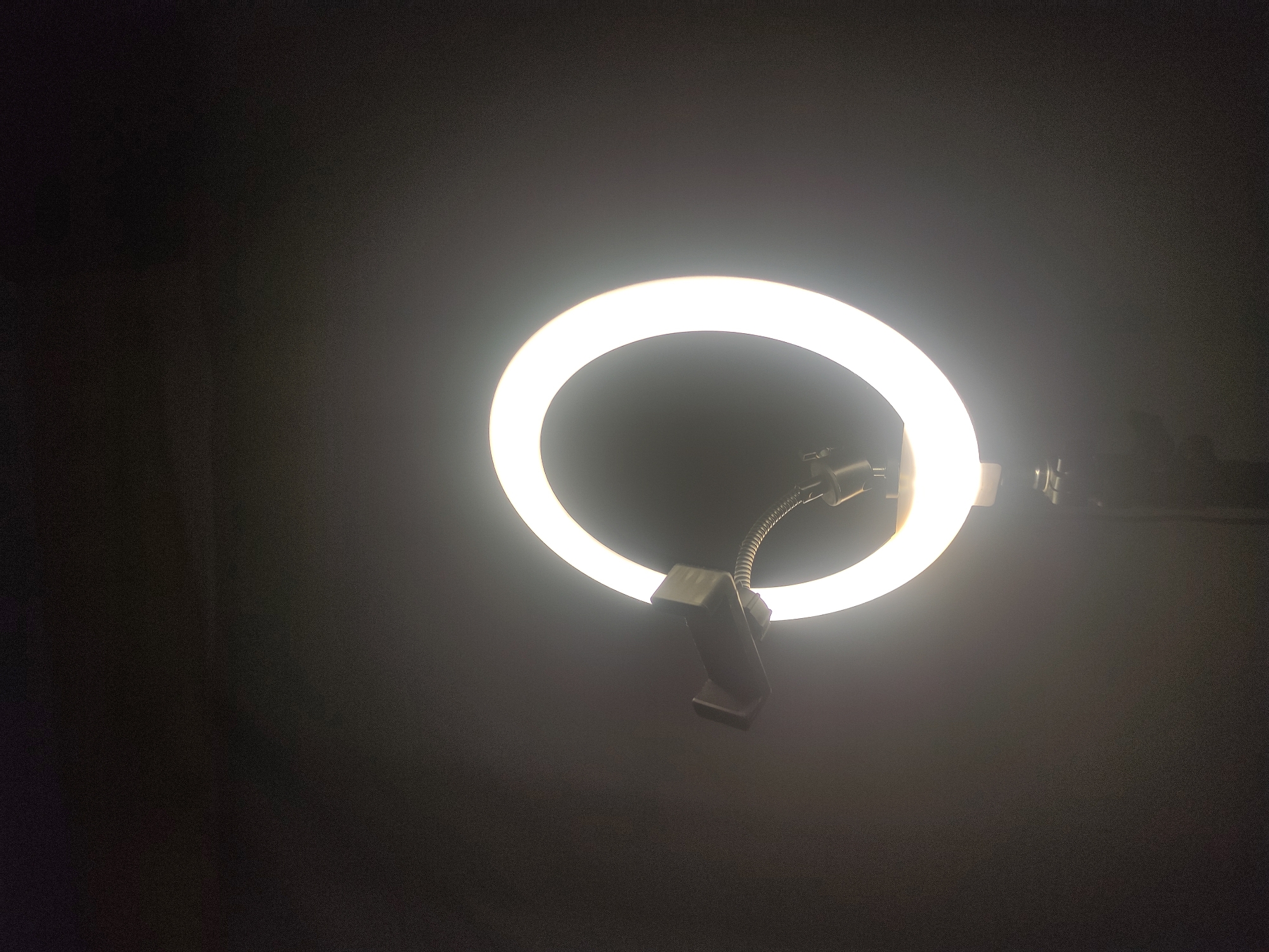 The image captures an illuminated LED ring light against a uniformly dark background. The circular light emits a bright, warm white glow. A black plastic smartphone holder, attached via a flexible metallic gooseneck arm, extends into the upper-left portion of the ring's interior; the holder is empty. Below the ring light, the upper section of a dark-colored tripod stand is visible, supporting the apparatus. The overall environment is sparsely lit, with the ring light serving as the primary illumination source. The scene is situated in the city of Neuheun, Indonesia.