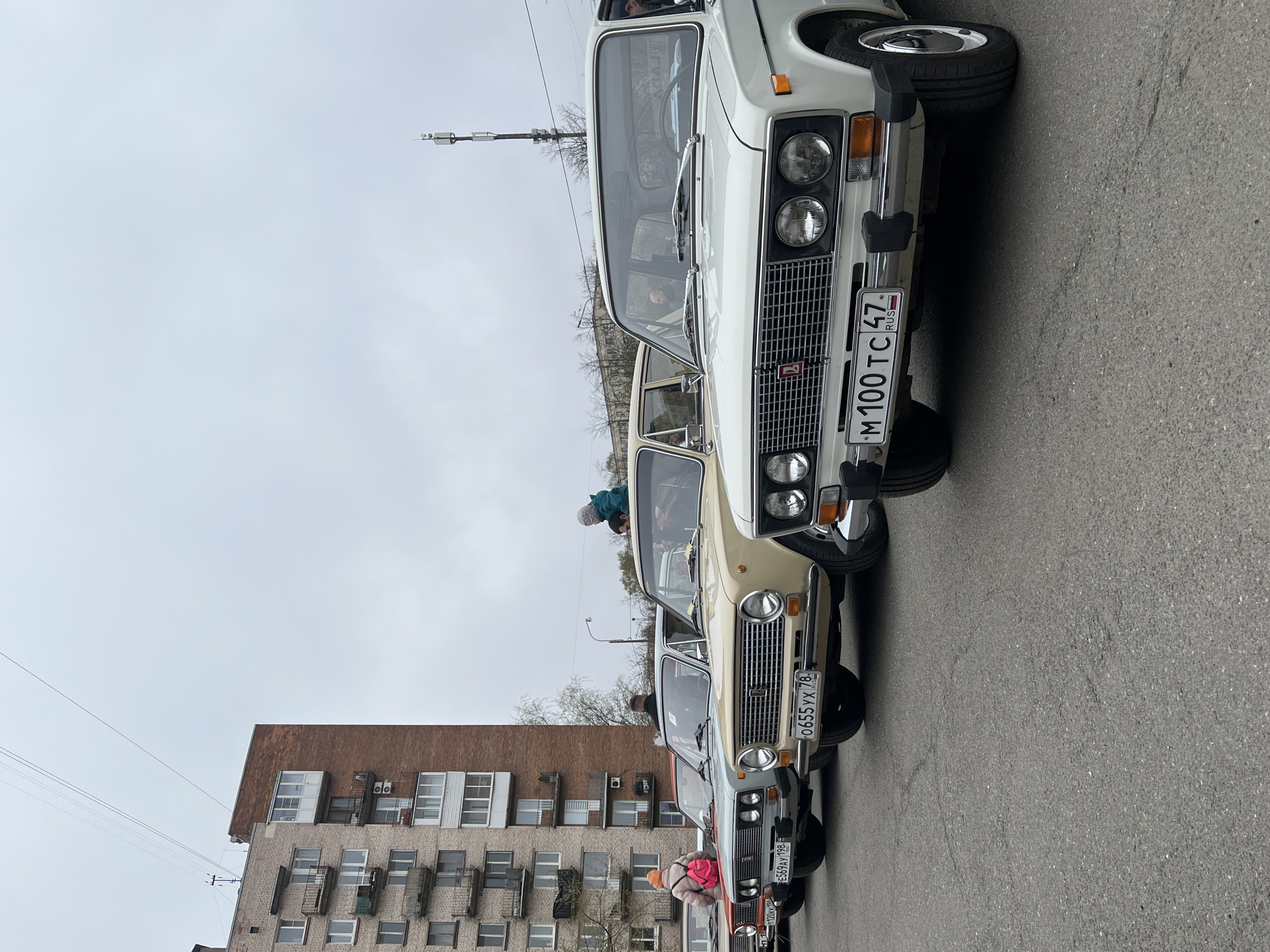 A collection of parked Lada cars, recognizable by their boxy shape and distinctive grille design, line a street in what appears to be Saint Petersburg, Russia. The cars are varying shades of white, beige, silver, and other colors. Each vehicle has a license plate, with some being distinctly visible. The license plate on the closest vehicle is marked "M 100 TC 47 RUS." Behind the cars is a brick apartment building, with many windows and a brown roof. On the street, several individuals can be seen around or inside the cars. The sky is overcast.