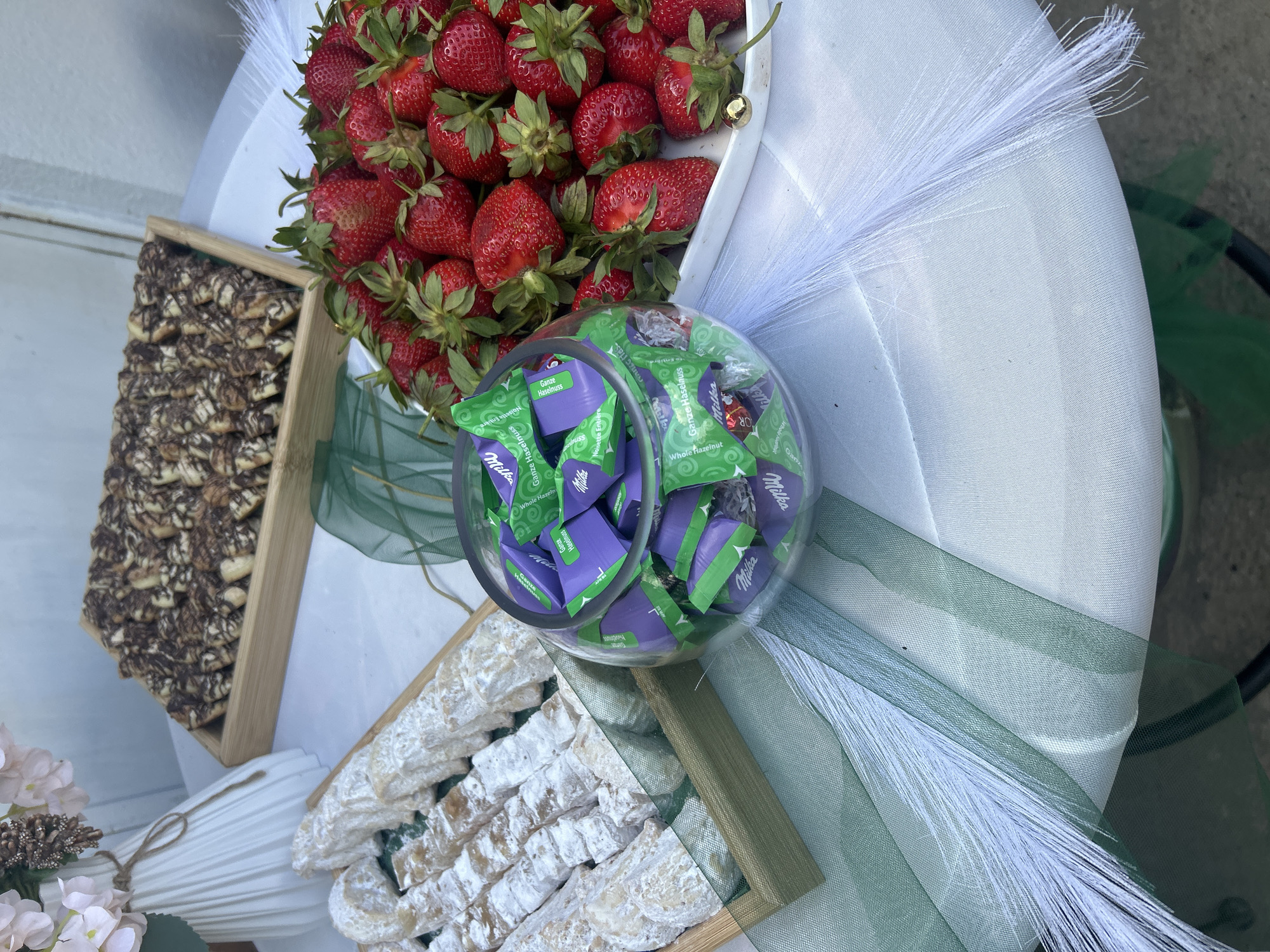 This eye-level shot depicts an assortment of food items arranged on a white surface. A bowl filled with Milka Whole Hazelnut chocolates, recognizable by their distinctive purple and green wrappers, takes center stage. Behind it is a white tray laden with fresh strawberries, showcasing their vibrant red color and green stems.
To the left and bottom of the frame are wooden trays containing baked goods. The tray on the left features treats with a marbled chocolate drizzle, while the tray on the bottom holds items generously dusted with powdered sugar. Green and white decorative elements are scattered around the food arrangement, adding an aesthetic touch.
The image originates from the city of Sandrovo, Bulgaria.