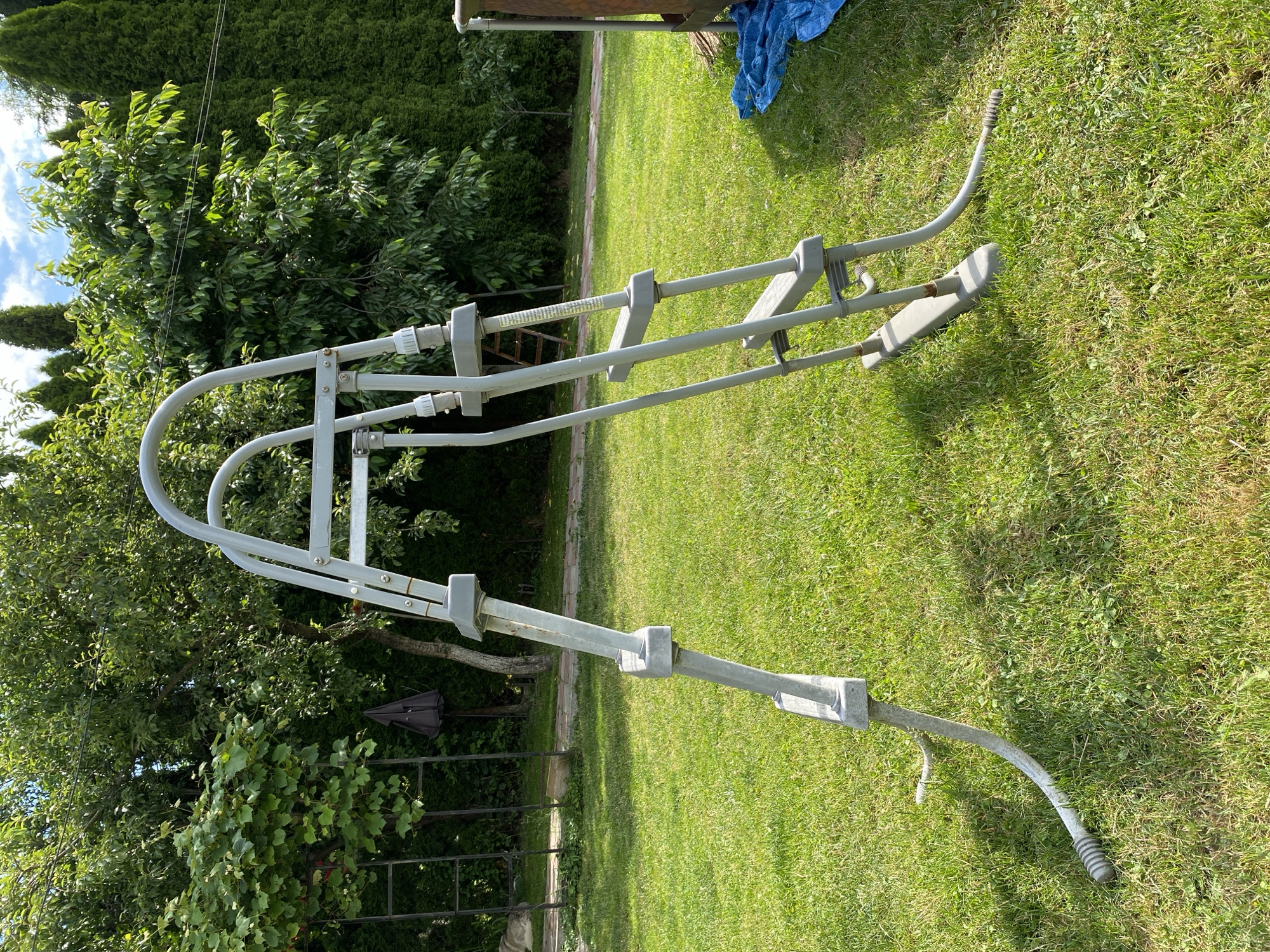 An overhead shot captures a gray metal pool ladder standing on a lush green lawn. The ladder, designed for an above-ground pool, has a U-shaped top and wide, flat steps. Its legs curve outwards, with rubber feet resting on the grass. Sunlight casts shadows from the ladder and trees onto the lawn. In the background, dense green foliage from various trees creates a natural backdrop, with a clear blue sky visible above. A dark umbrella and some gardening equipment are partially visible to the left. A blue tarp lies on the grass to the right, near a metal structure.