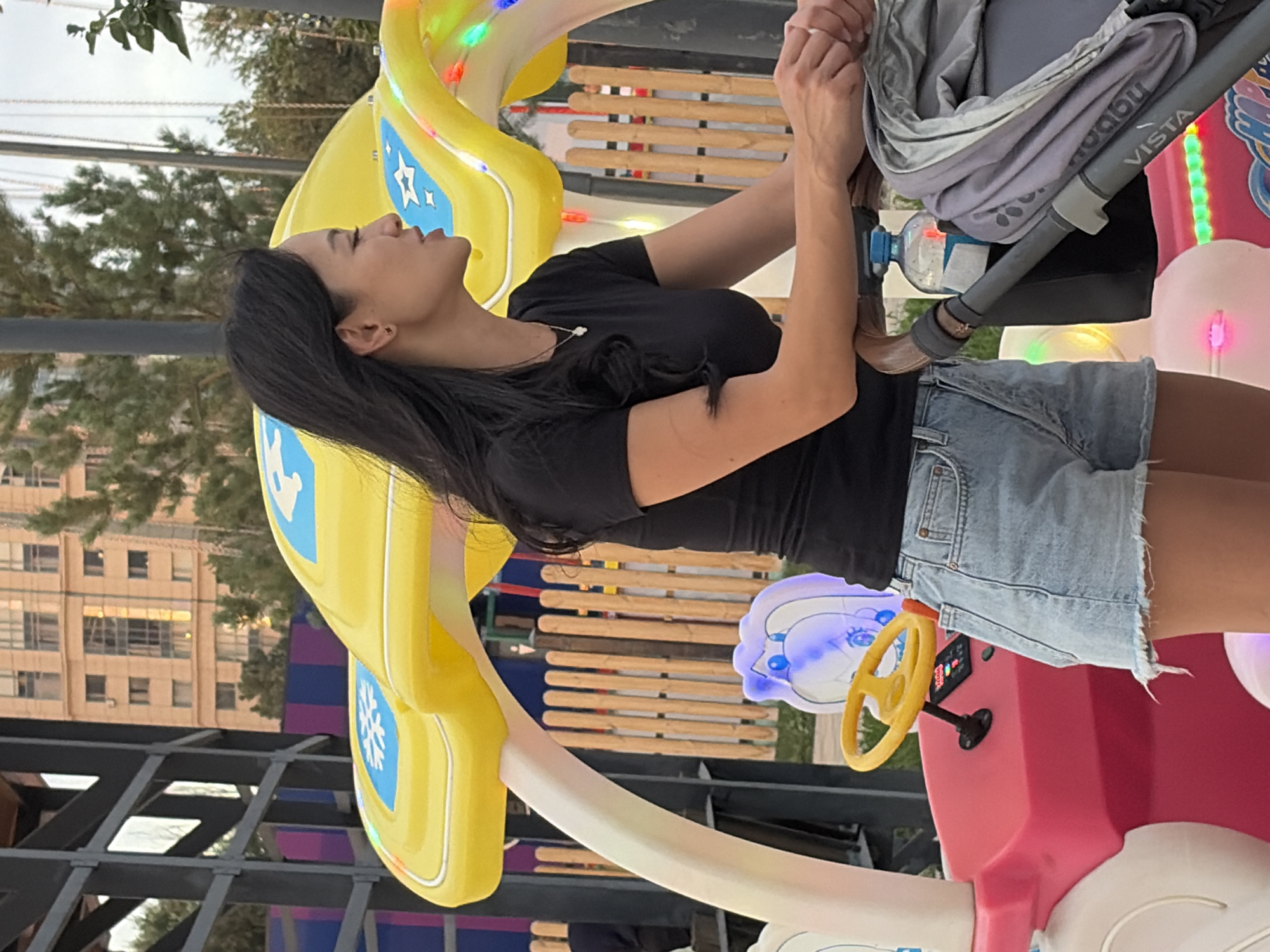 In an outdoor amusement park setting, a woman with long dark hair, wearing a black t-shirt and denim shorts, stands next to a brightly colored children's ride. The ride is yellow and pink, with a yellow steering wheel. In front of the woman is a stroller with a gray cover. The background shows a building, trees, and a wooden fence. The lighting suggests it is daytime, with some artificial lights on the ride.