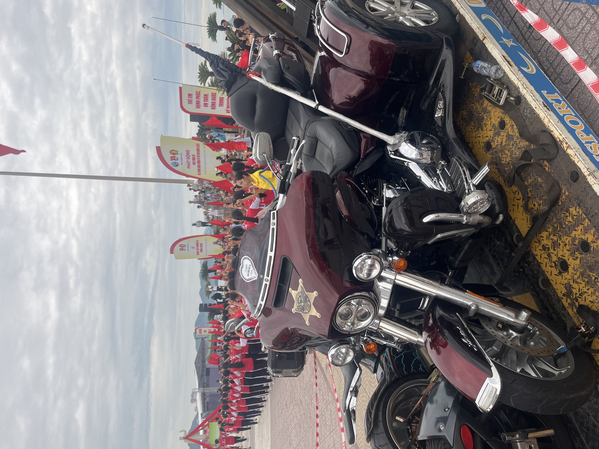 The image shows a close-up view of two maroon and black Harley Davidson-style motorcycles, positioned on what appears to be a transport platform or truck bed. The motorcycle in the foreground has a prominent "Sheriff" style star badge on its windshield. In the background, a large crowd of people wearing red and white attire is visible. Banners with Vietnamese text and symbols are also present. A partial view of the sky and a few palm trees can be seen in the far background. The OCR text mentions “Nha Trang Ward, Vietnam”, which, combined with visual elements, suggests the location of the image.